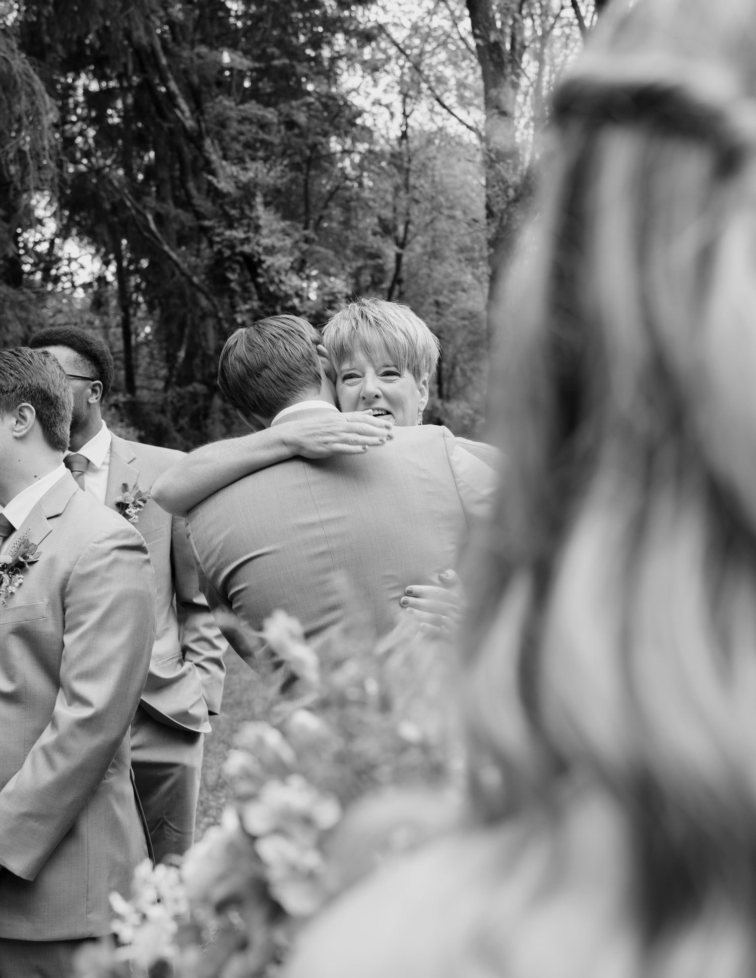Black and white photo of woman warmly embracing groom at outdoor wedding ceremony.