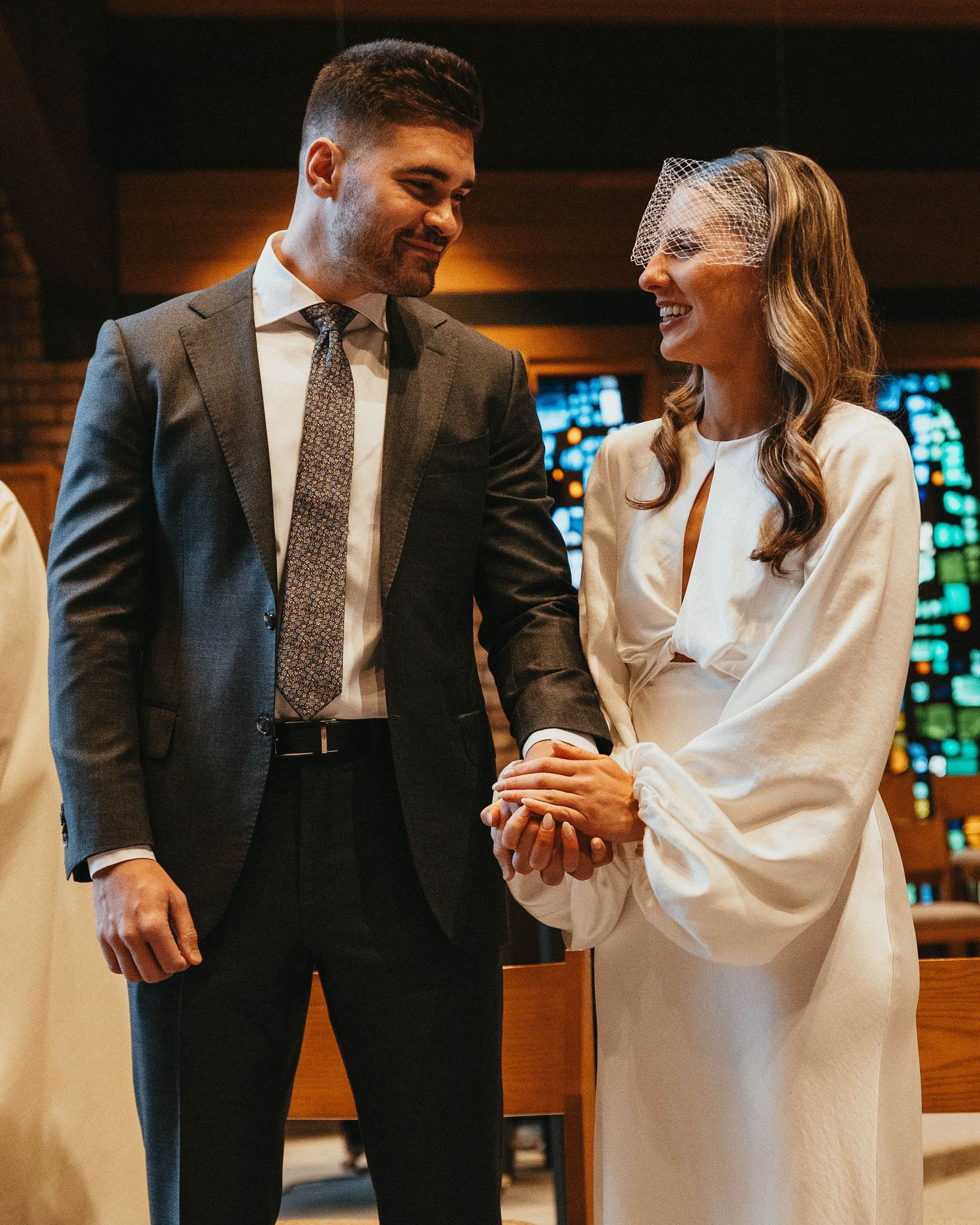 Bride and groom hold hands and smile at each other during their wedding ceremony inside a church.