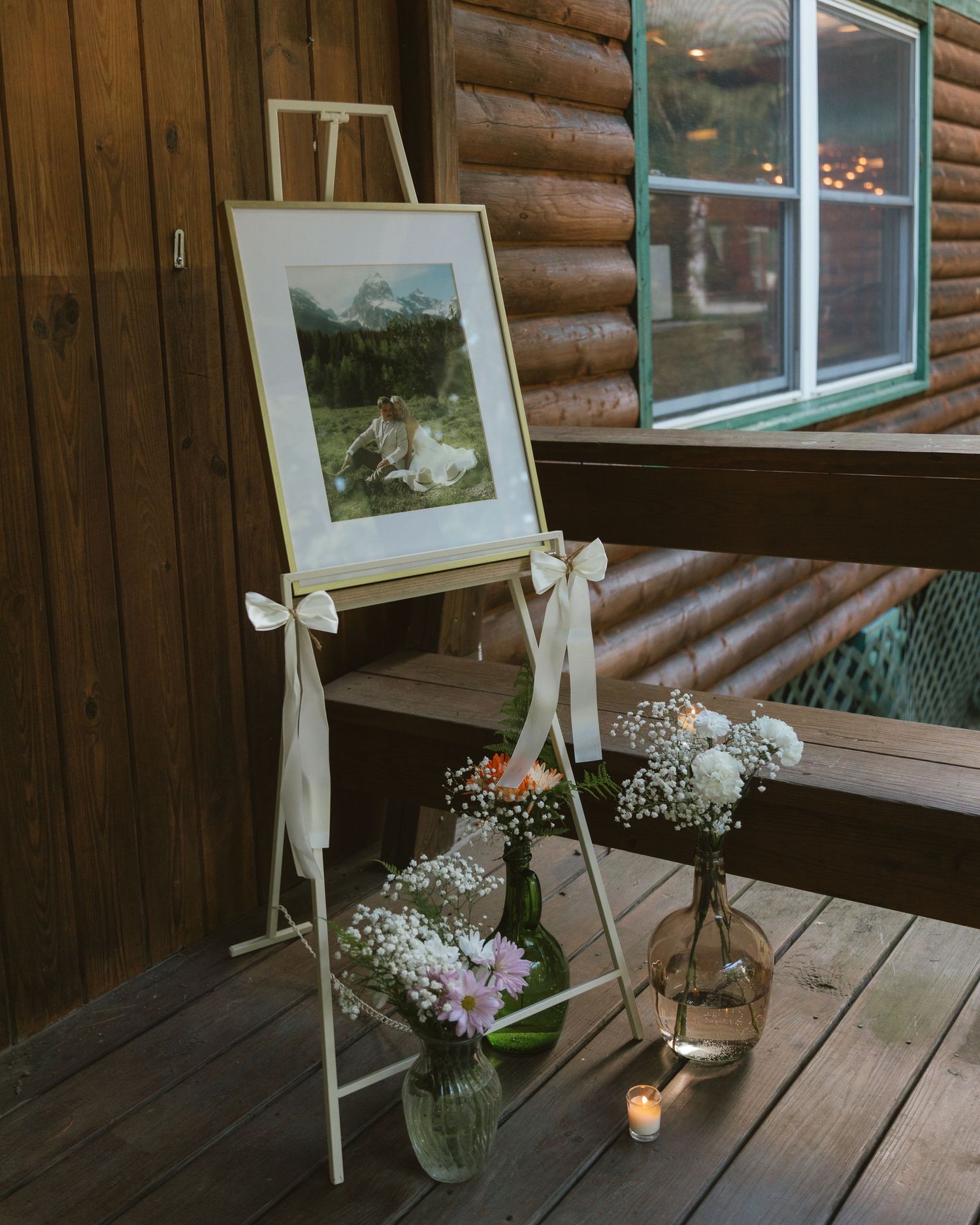 Elegant wedding easel with framed photo, white bows, and floral arrangements on a rustic log cabin porch.