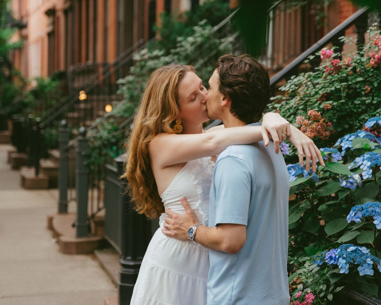 Couple sharing a romantic kiss on a charming city sidewalk lined with blooming hydrangeas and brownstone buildings.