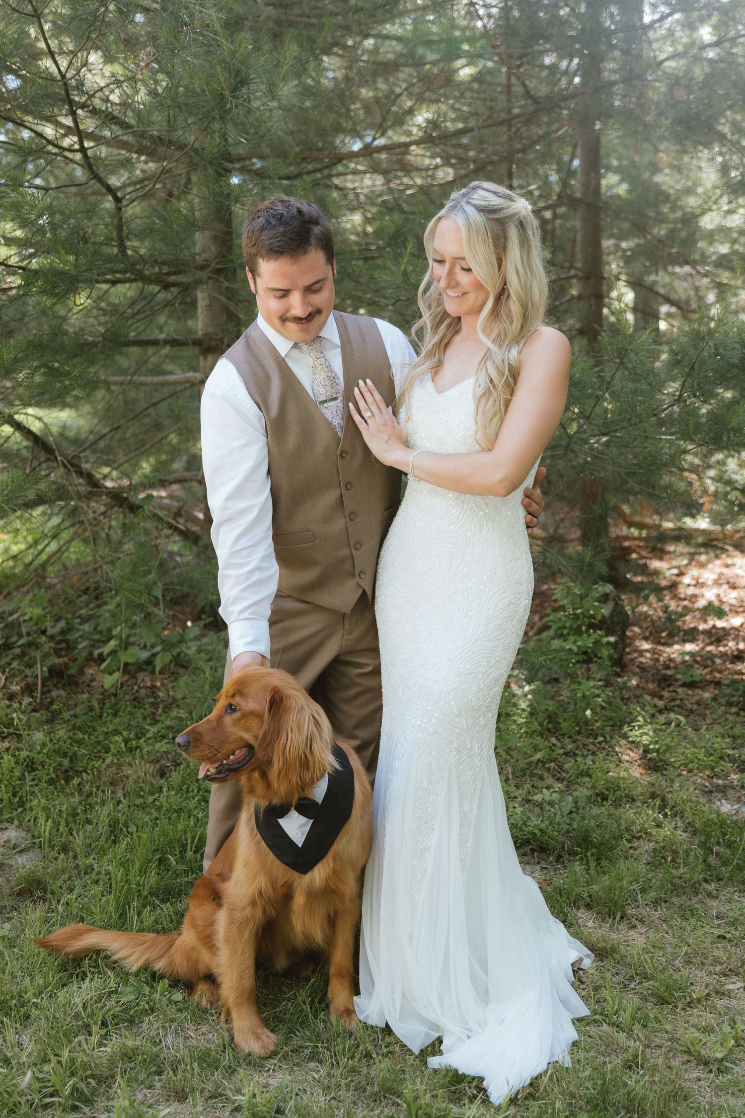Newlyweds in wedding attire smile while groom pets their golden retriever dog wearing a tuxedo bandana.