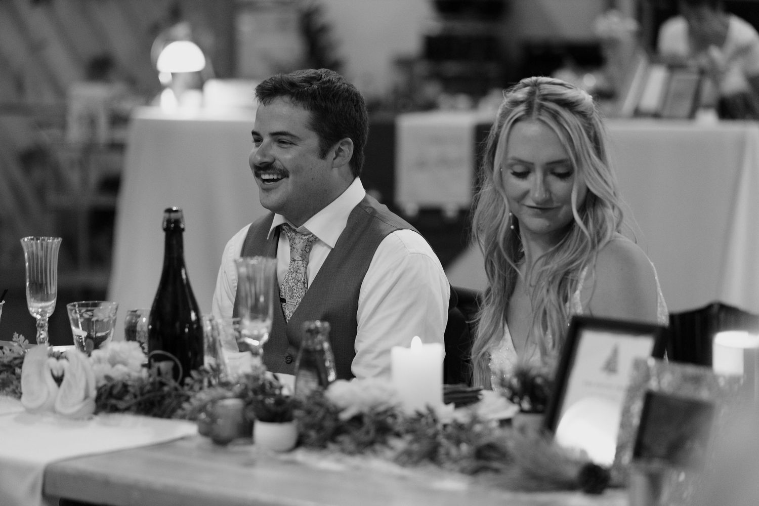 Black and white photo of smiling bride and groom seated at wedding reception head table with candles and flowers.