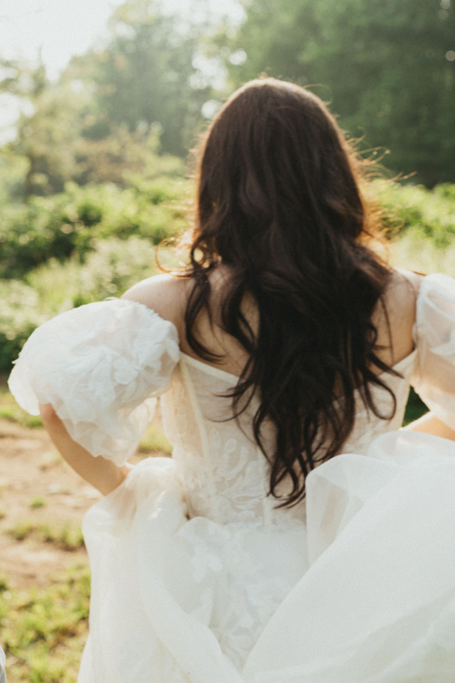 Bride with long dark wavy hair in white off-shoulder lace wedding dress walking through sunlit garden.