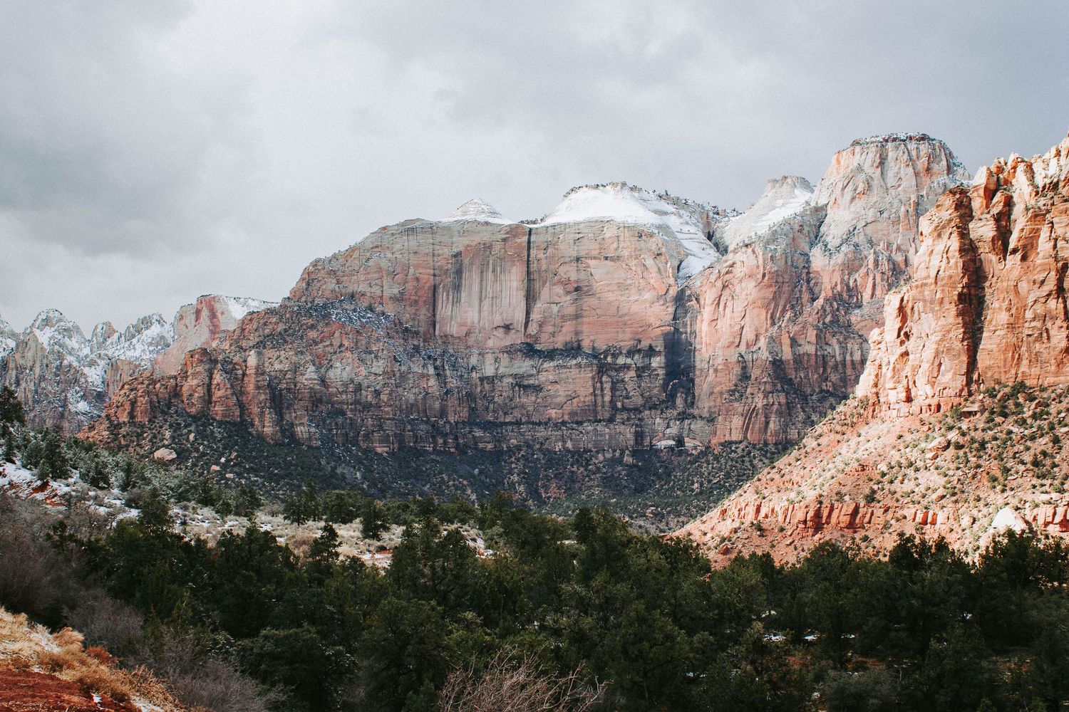 Snow-dusted red rock cliffs and pine trees under cloudy skies in Zion National Park, Utah.