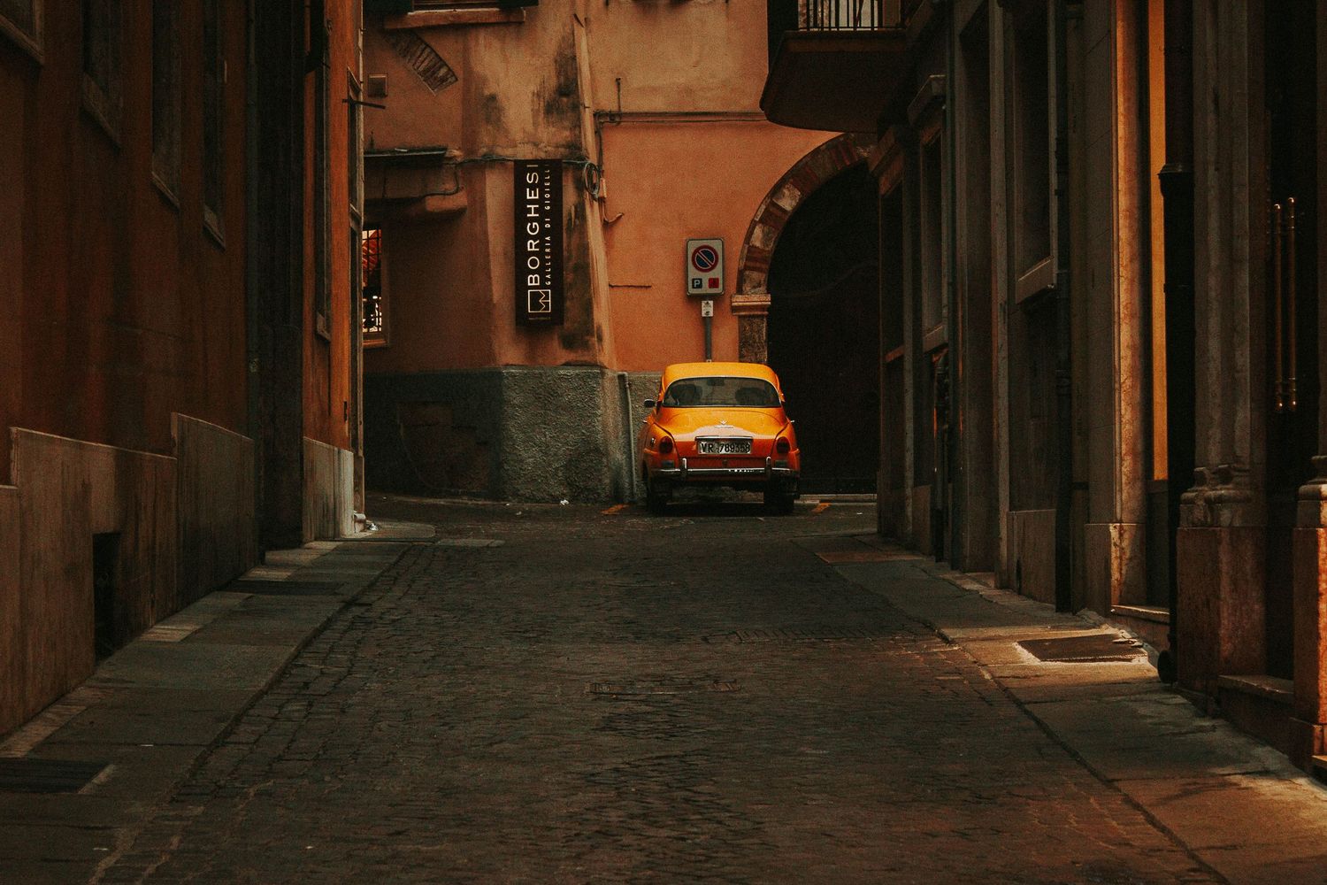 A vintage orange car parked in a narrow cobblestone alley between aged terracotta buildings in Italy.