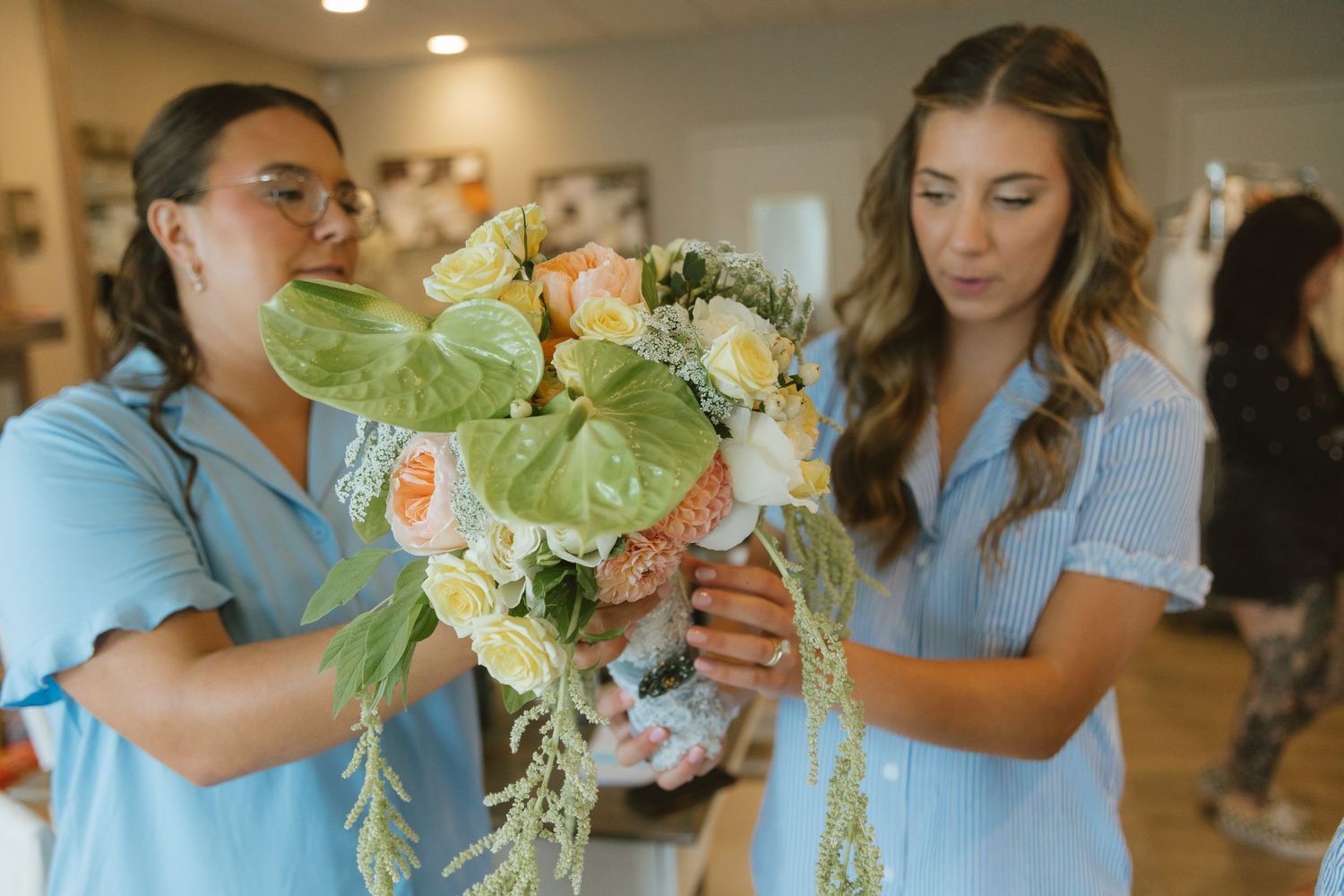 Two women in blue shirts arrange a beautiful pastel floral bouquet with roses and tropical leaves in a bright studio.