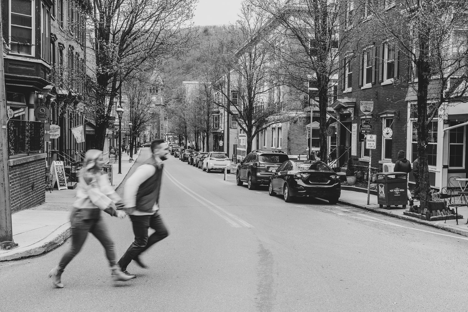 Candid black and white photo of an engaged couple holding hands and running across a street, an artistic engagement session in Jim Thorpe.