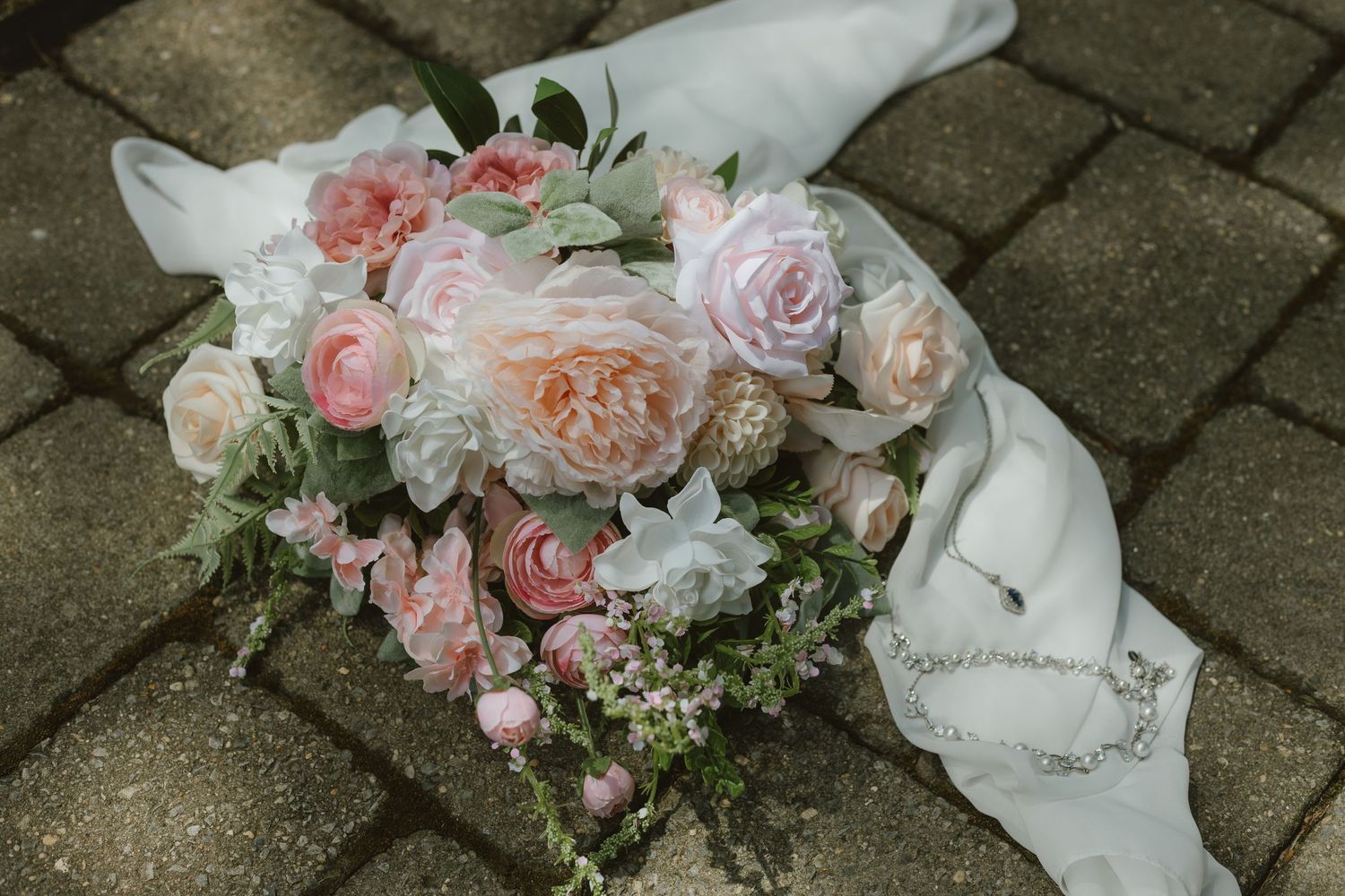 A romantic bridal bouquet of pink and peach roses with greenery rests on cobblestone beside a white veil and pearl necklace.