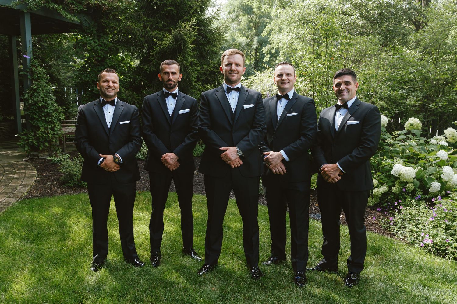 Five groomsmen in black tuxedos with bow ties pose together outdoors in a lush garden setting.