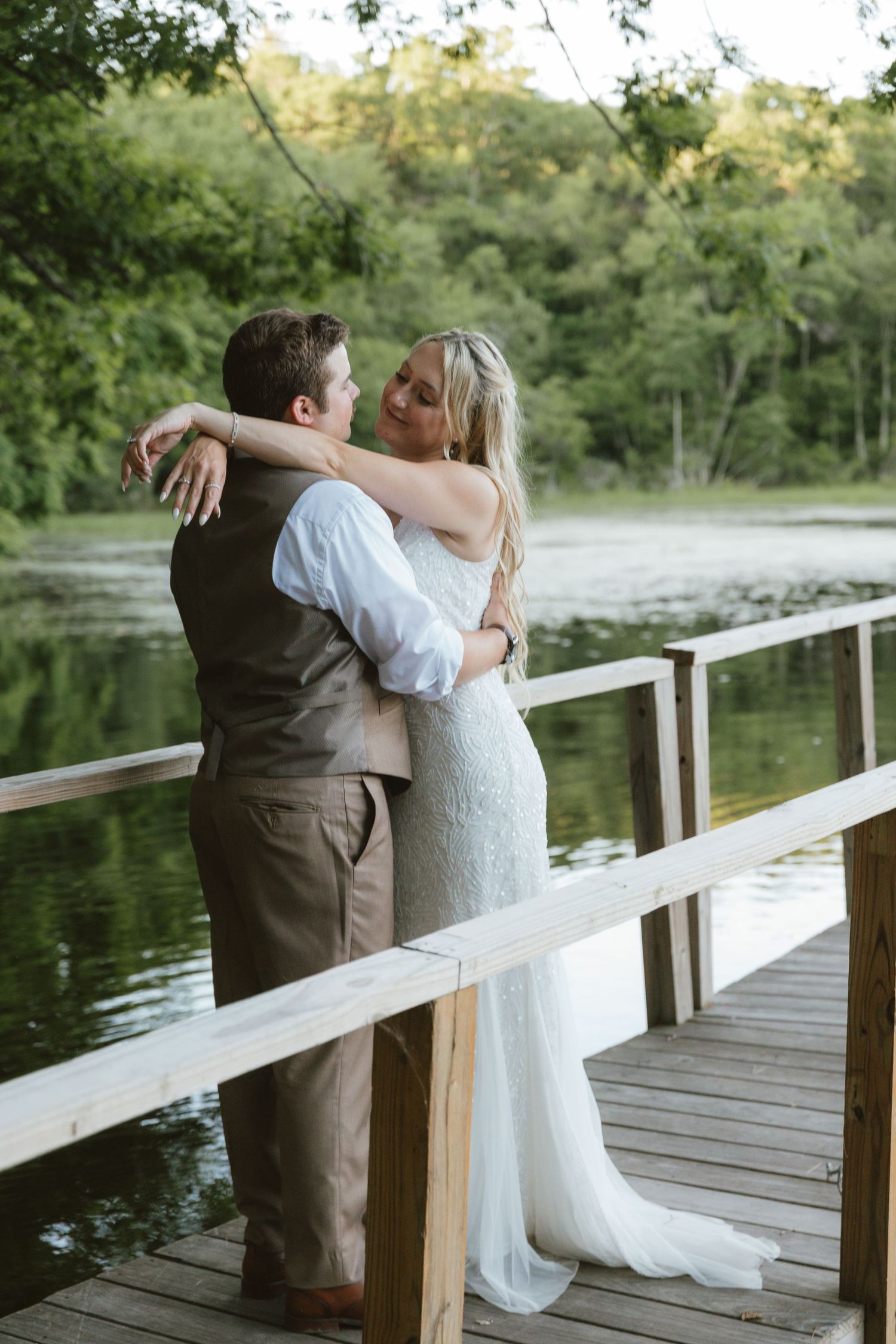 Bride and groom embracing on a wooden dock by a serene lake surrounded by lush green trees.
