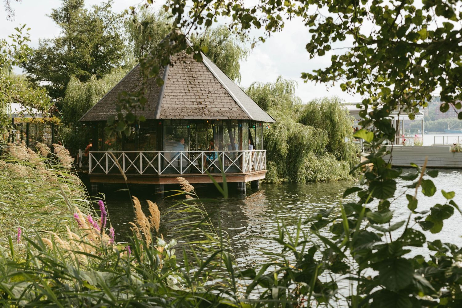 A charming wooden gazebo on a tranquil pond surrounded by weeping willows and lush greenery.