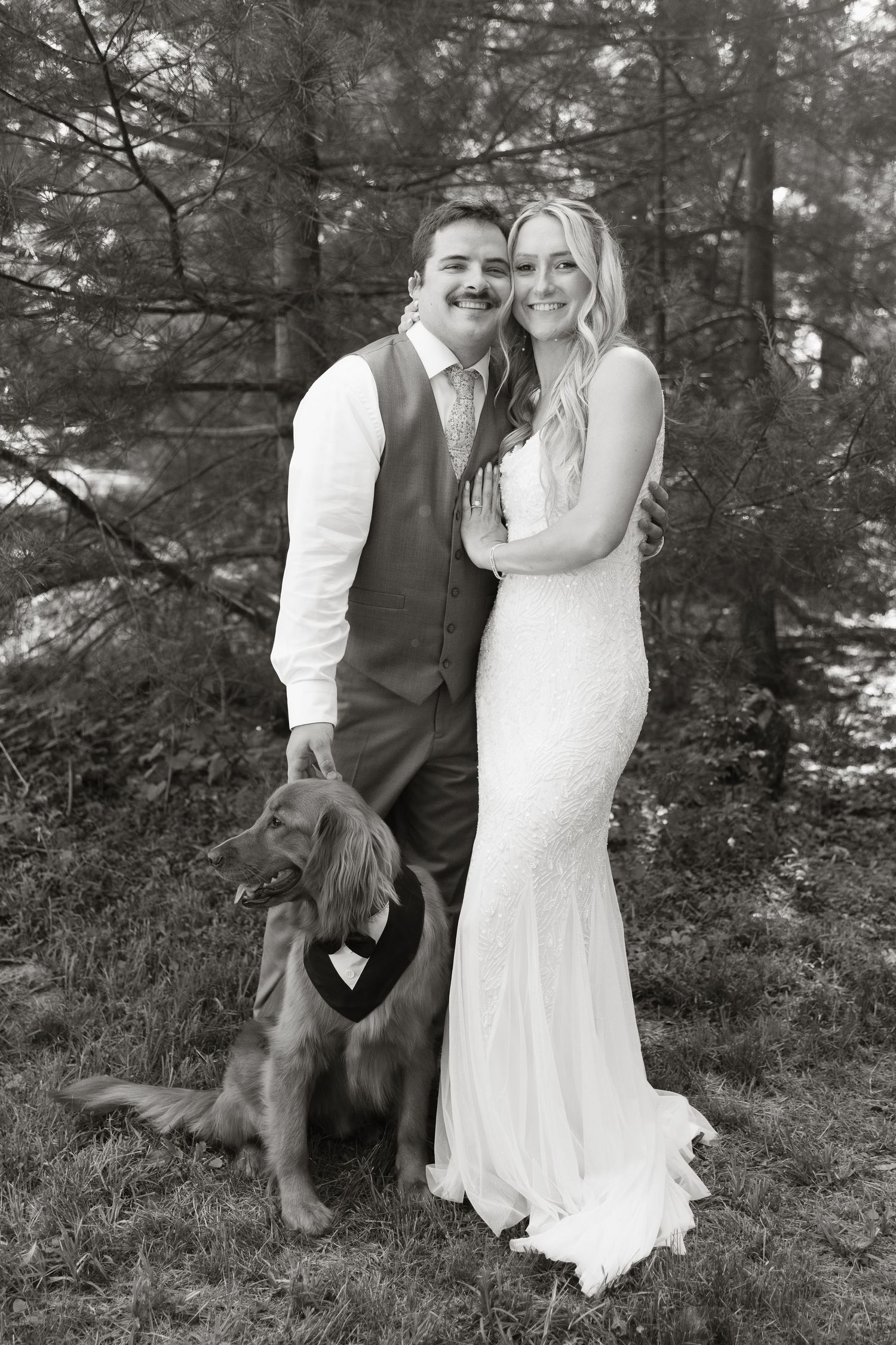Black and white wedding photo of smiling bride and groom posing outdoors with their golden retriever dog wearing a bow tie.