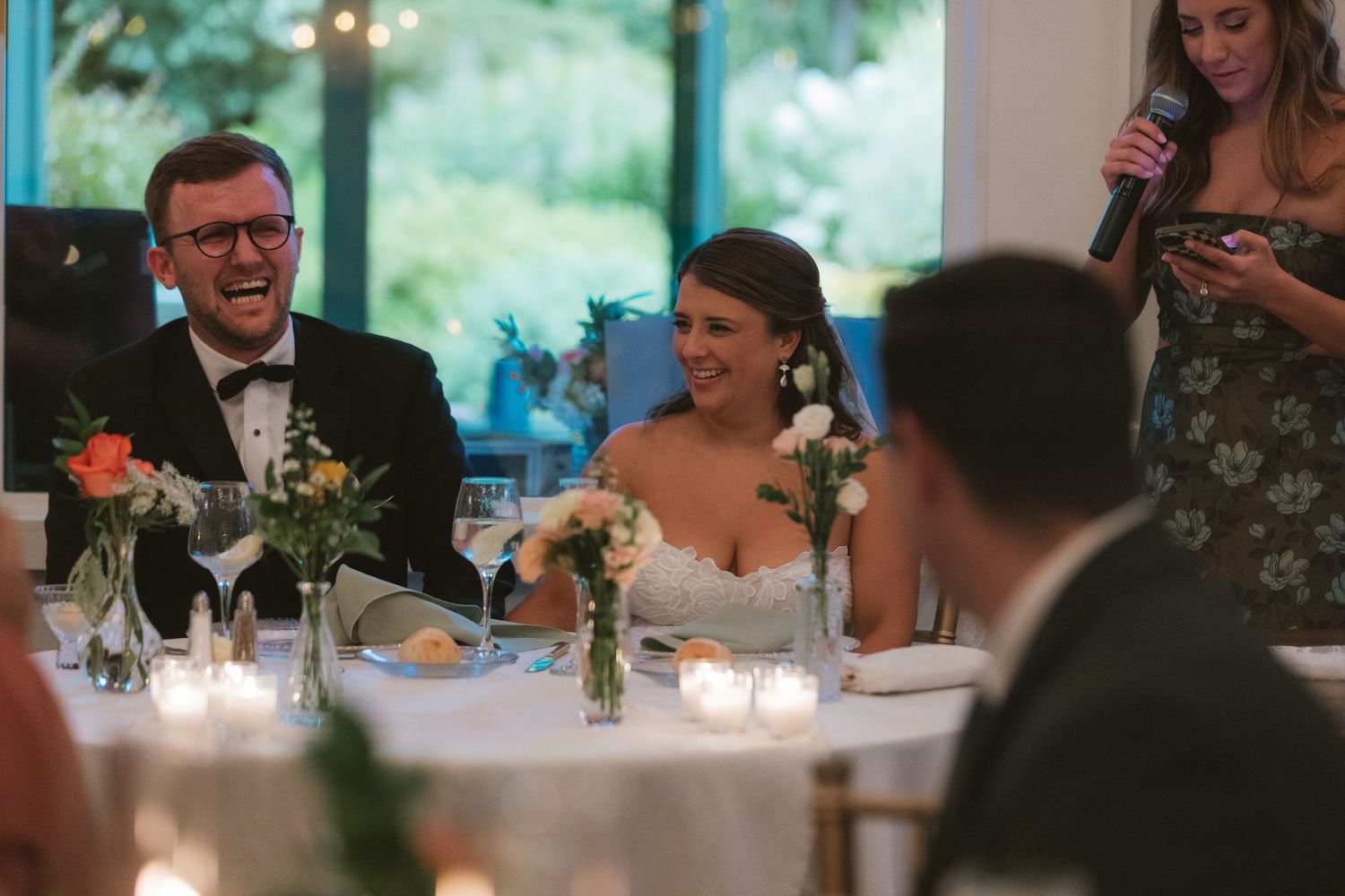 Bride and groom laughing joyfully at wedding reception table while bridesmaid gives speech with microphone.