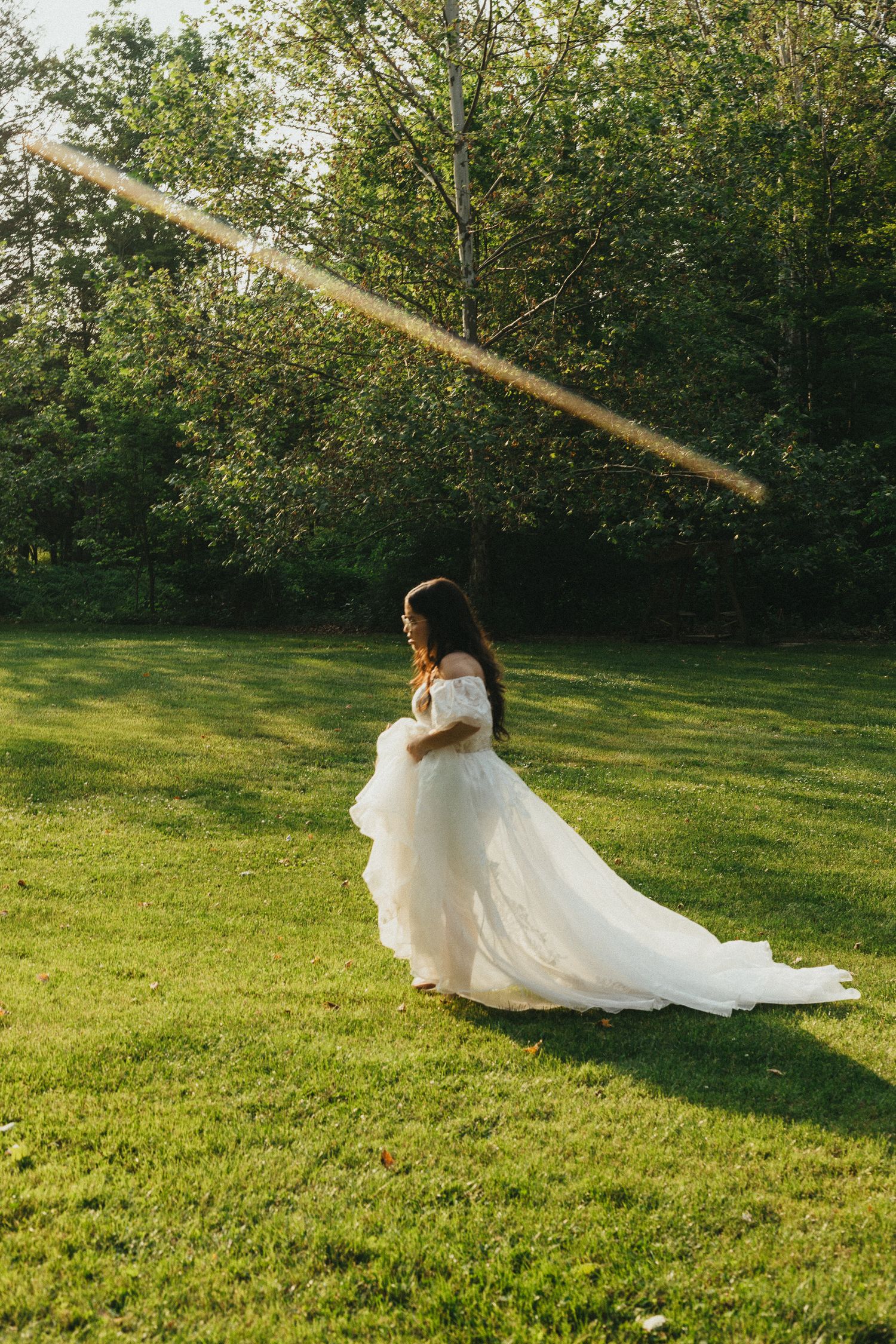 Bride in off-shoulder ball gown twirling on green lawn with sunlit trees in background.