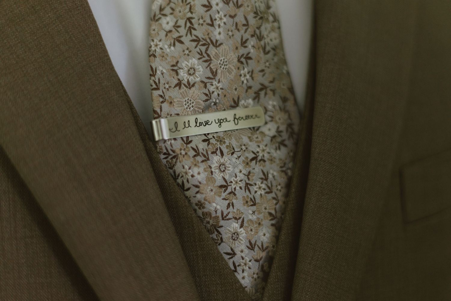 Close-up of a floral tie with a personalized silver tie clip engraved with a message on a olive suit jacket.