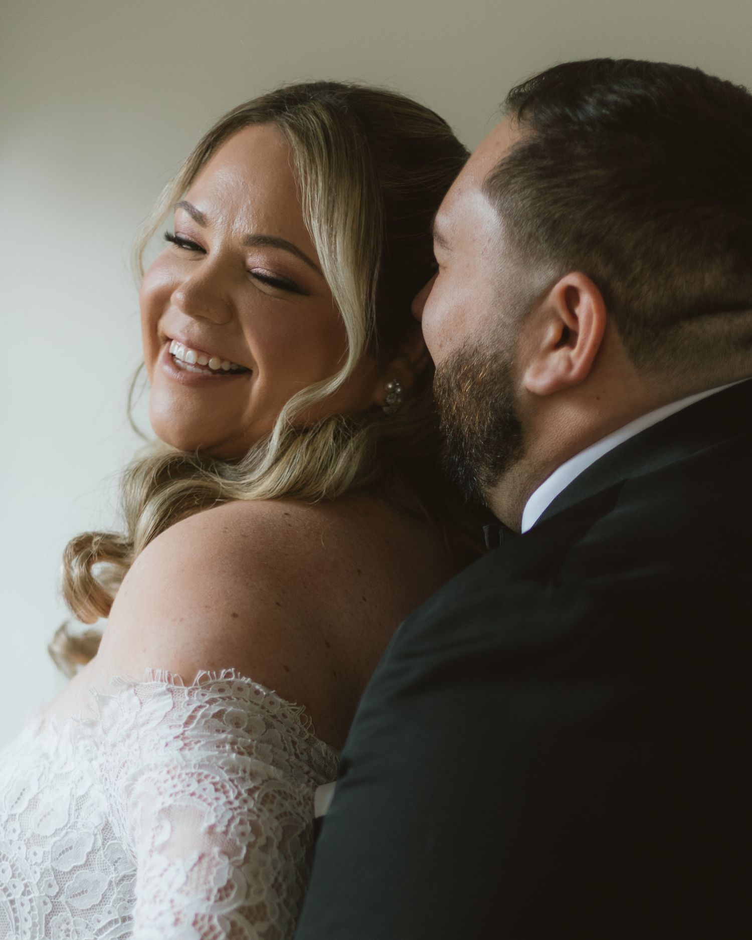 A smiling bride in an off-shoulder lace gown shares a tender moment with a bearded groom in a dark suit.