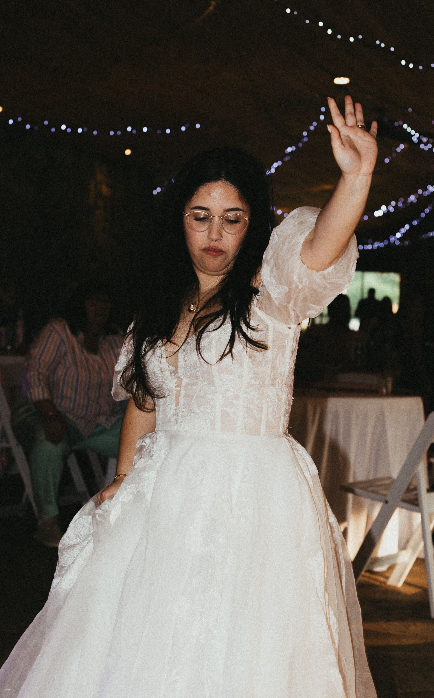 Bride in white lace gown dancing with arm raised at indoor wedding reception with string lights.