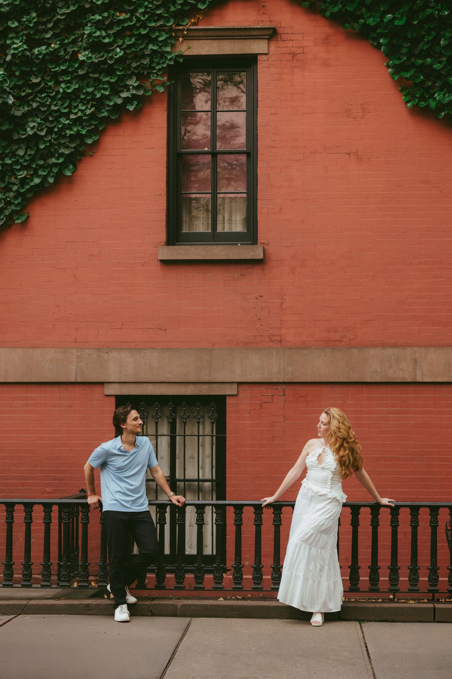 A couple stands apart on a sidewalk railing before a red brick building with ivy, the woman in a white dress, man in blue shirt.