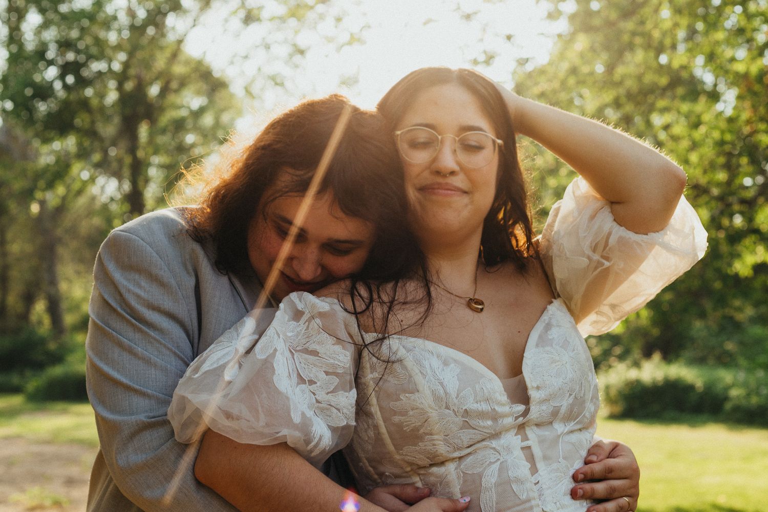 Couple sharing tender embrace in golden sunlight at outdoor wedding, groom nuzzling bride in white dress.
