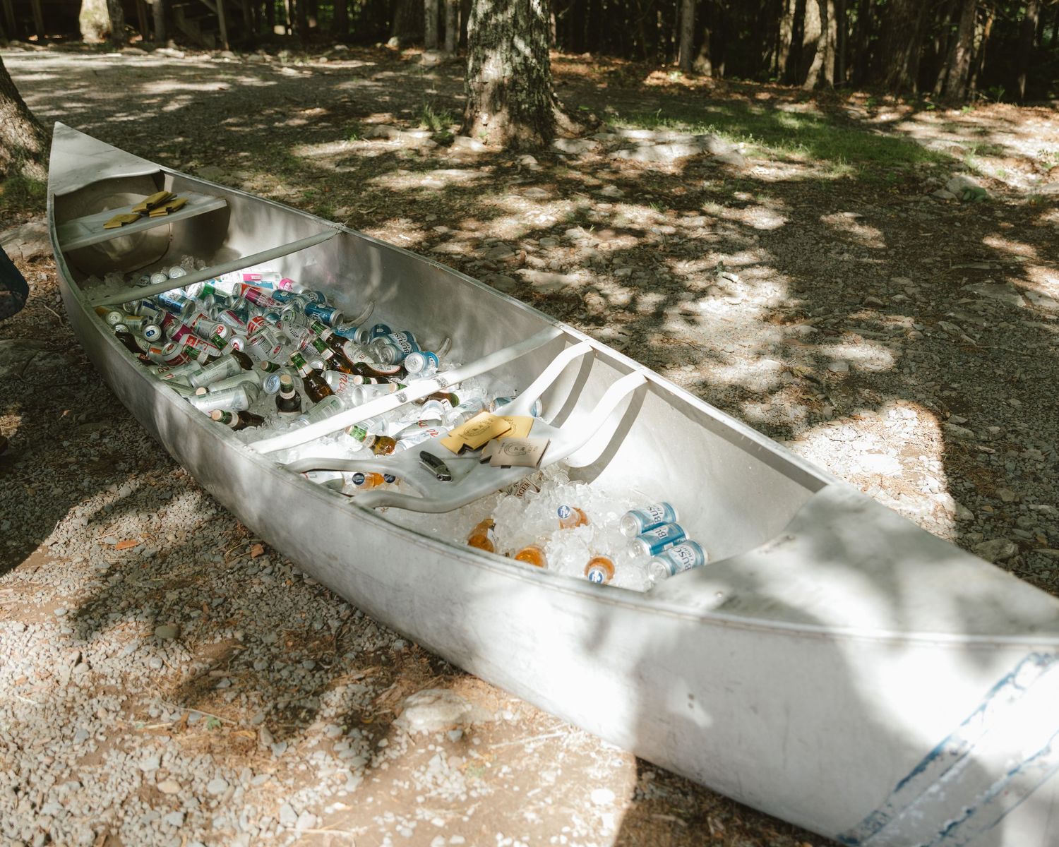 Silver canoe filled with beverages and snacks used as a cooler, placed on shaded ground surrounded by trees.