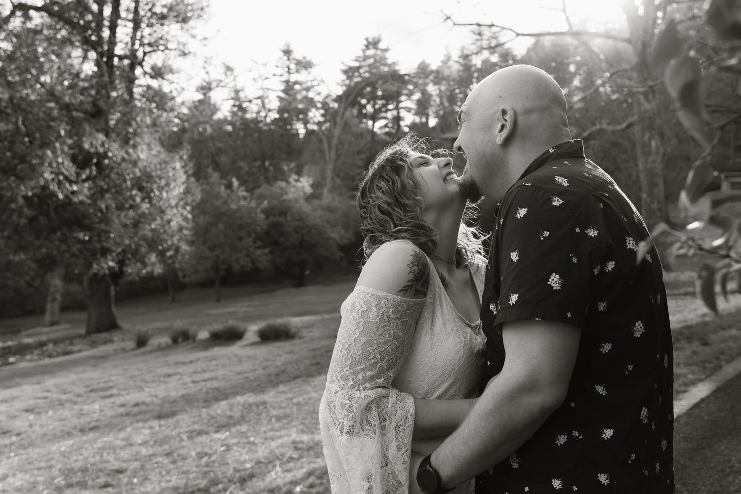 Black and white photo of a couple sharing a romantic kiss outdoors at their wedding ceremony surrounded by trees.