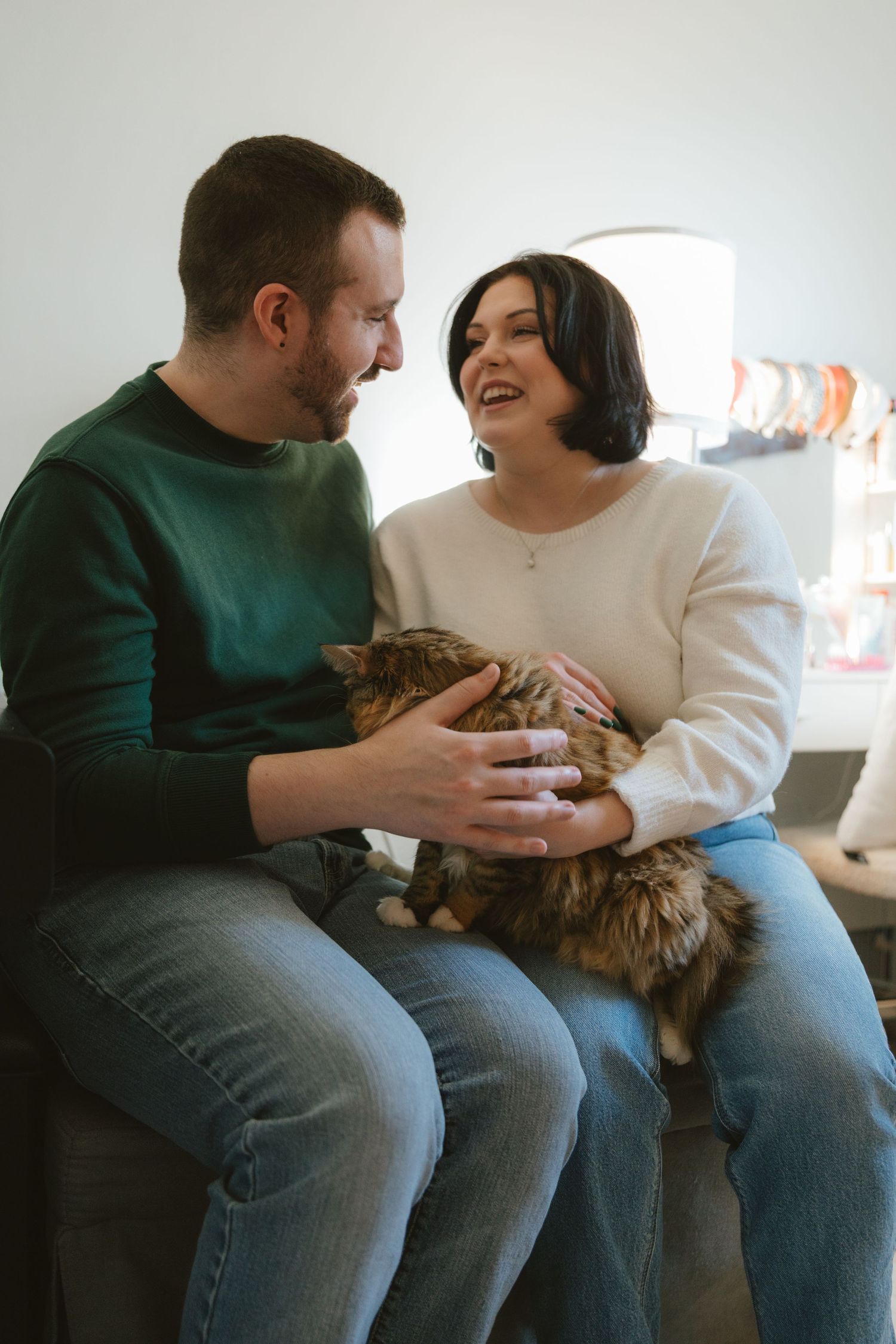 A happy couple sitting together, smiling and petting a tabby cat in a bright living room.
