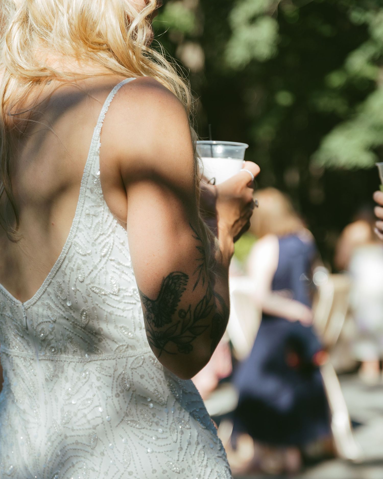 Close-up of a tattooed bride in a white dress holding a drink at an outdoor wedding.