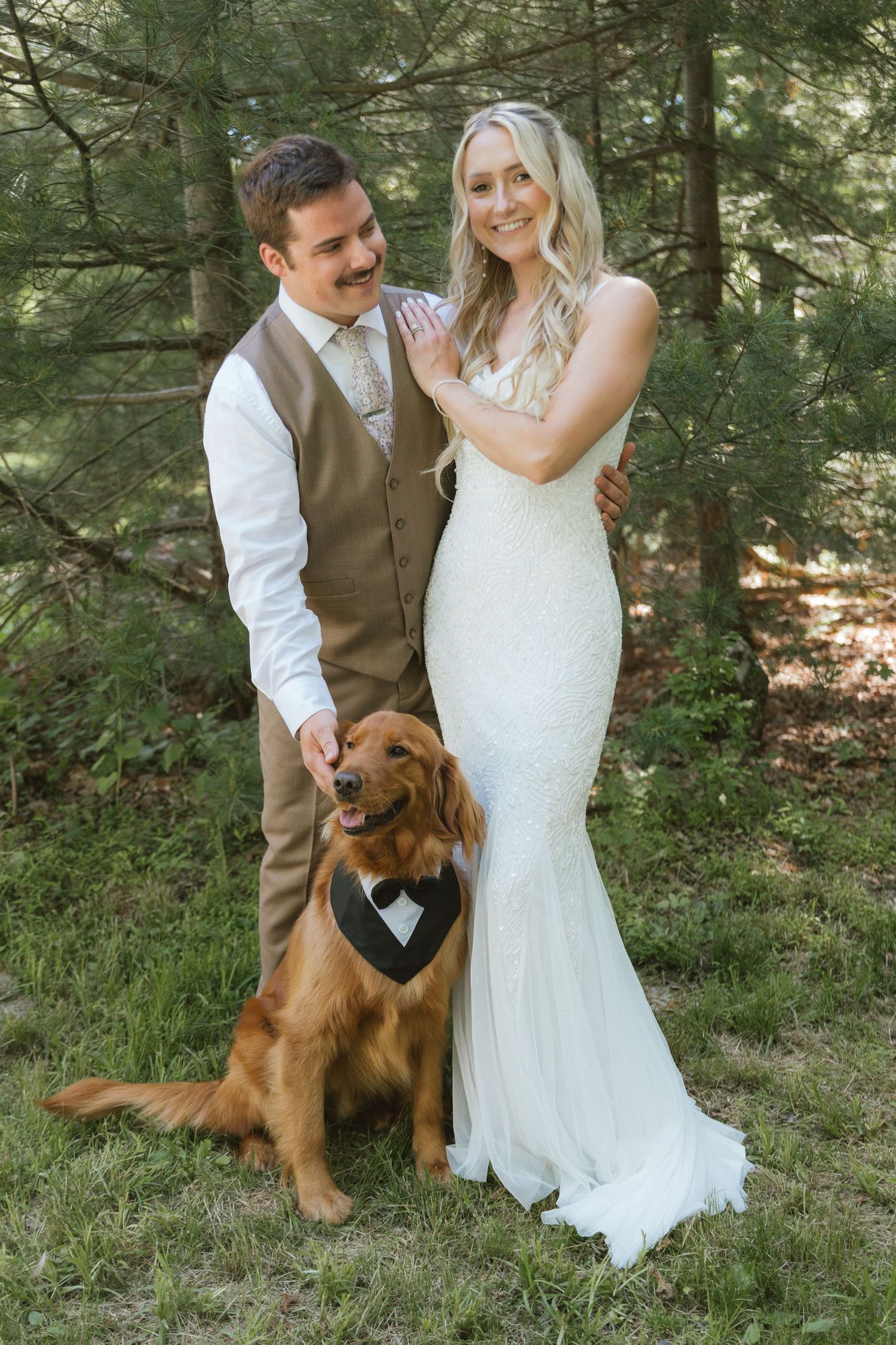 Smiling bride and groom in wedding attire pose outdoors with their golden retriever wearing a bow tie sign.