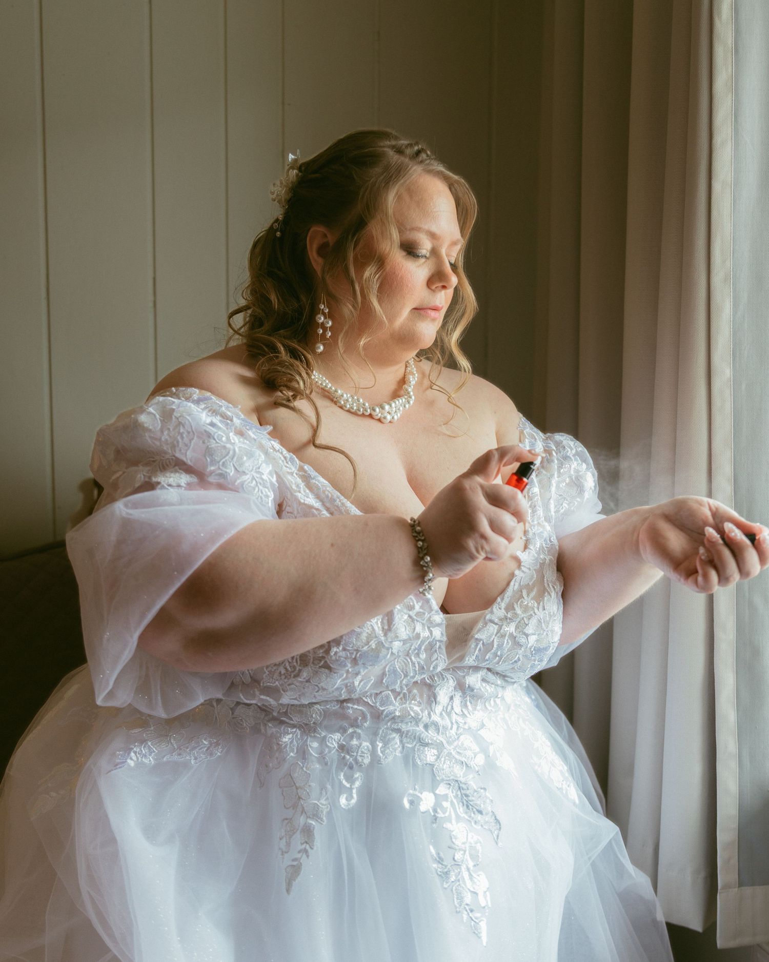 A plus-size bride in a white lace ball gown sprays perfume on her wrist near a window on her wedding day.