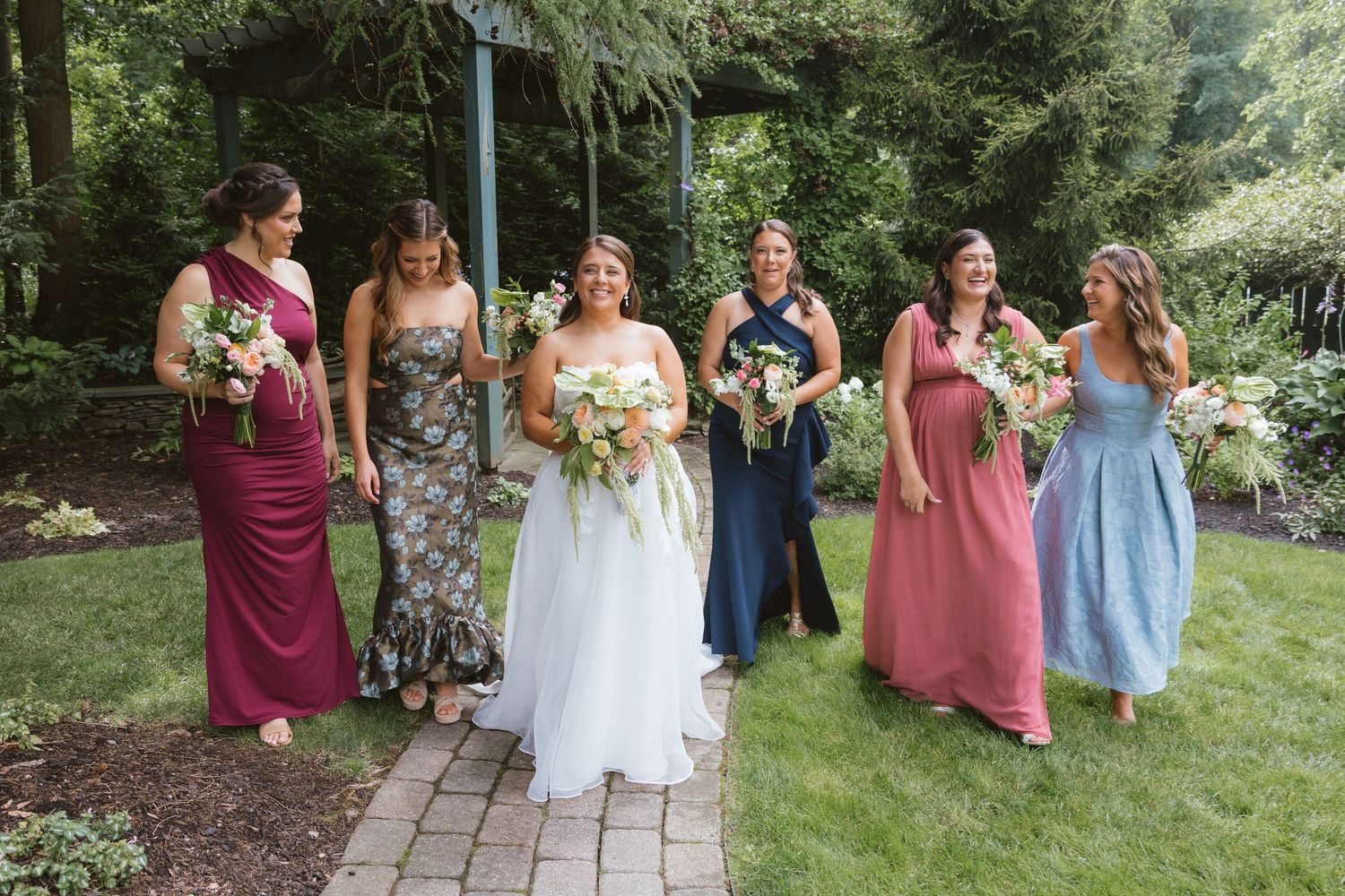 Bride in white gown walks with five bridesmaids in colorful mismatched dresses on a garden path.