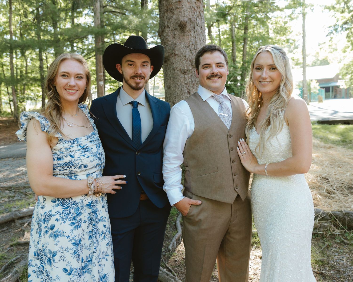 Two couples pose outdoors among pine trees, one bride in white, another in floral dress, grooms in suit and cowboy hat.