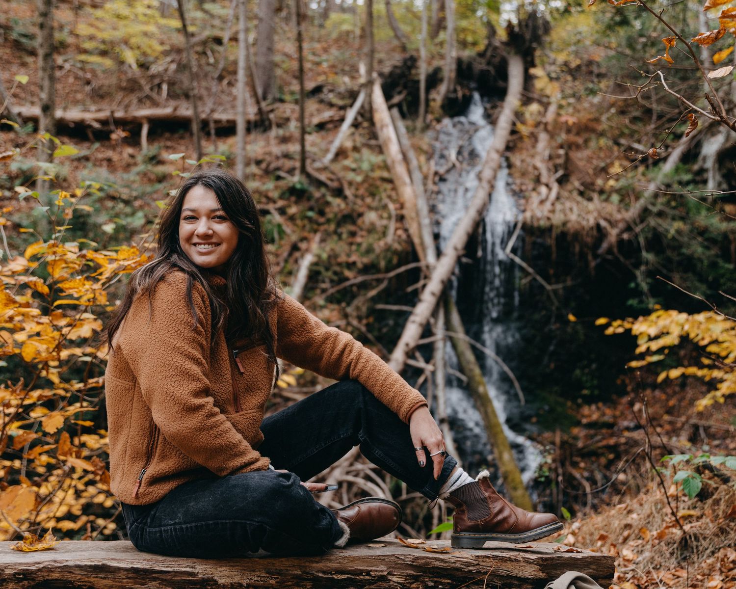 Woman in brown sweater smiling on a log with autumn foliage and waterfall backdrop.