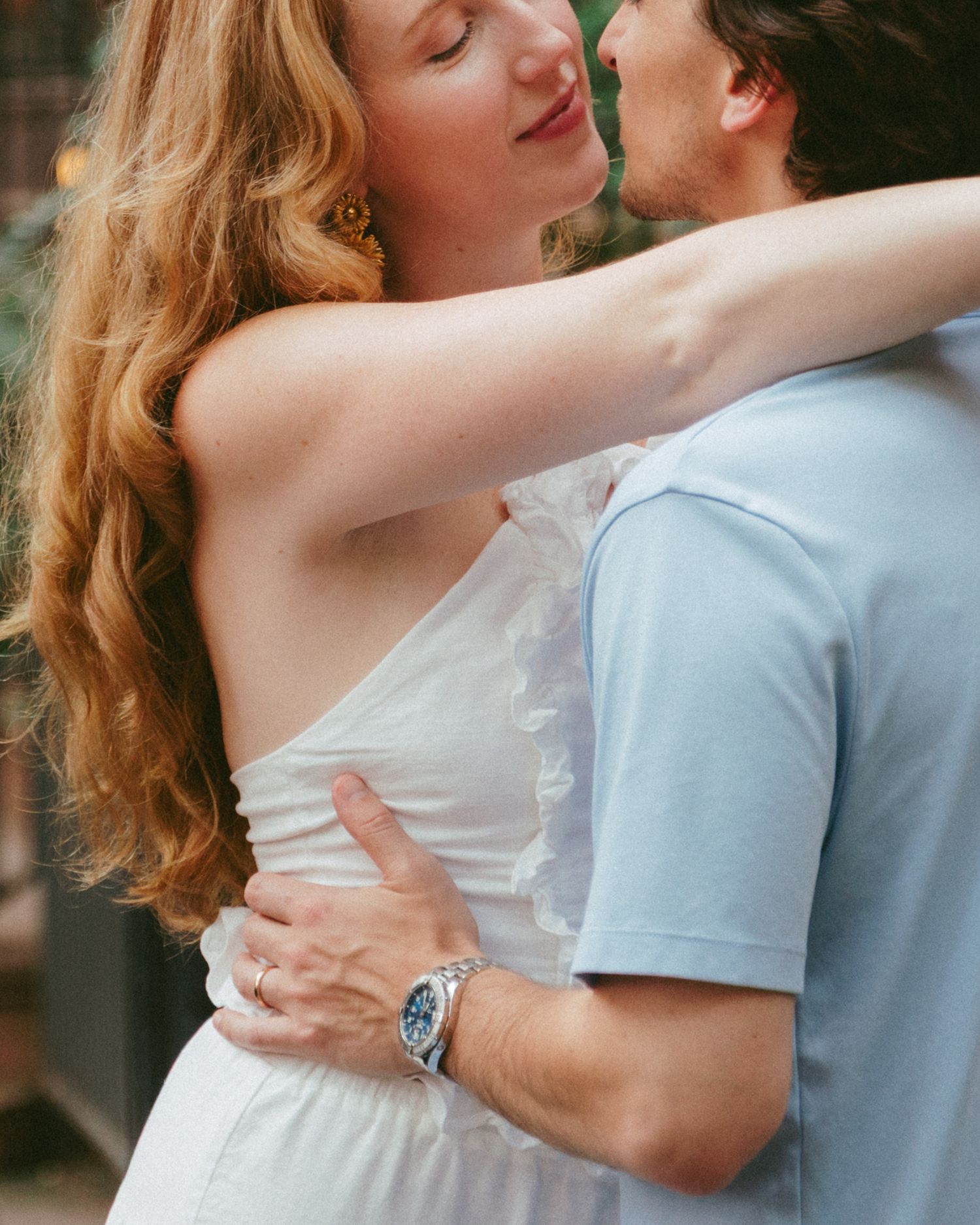 A couple sharing a romantic embrace outdoors, woman in white dress with long red hair hugging a man in a light blue shirt.