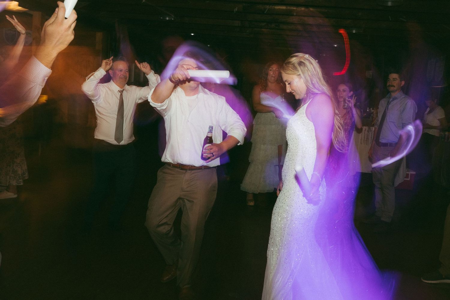 Bride in white gown and guests dance under purple lights during lively wedding reception celebration.