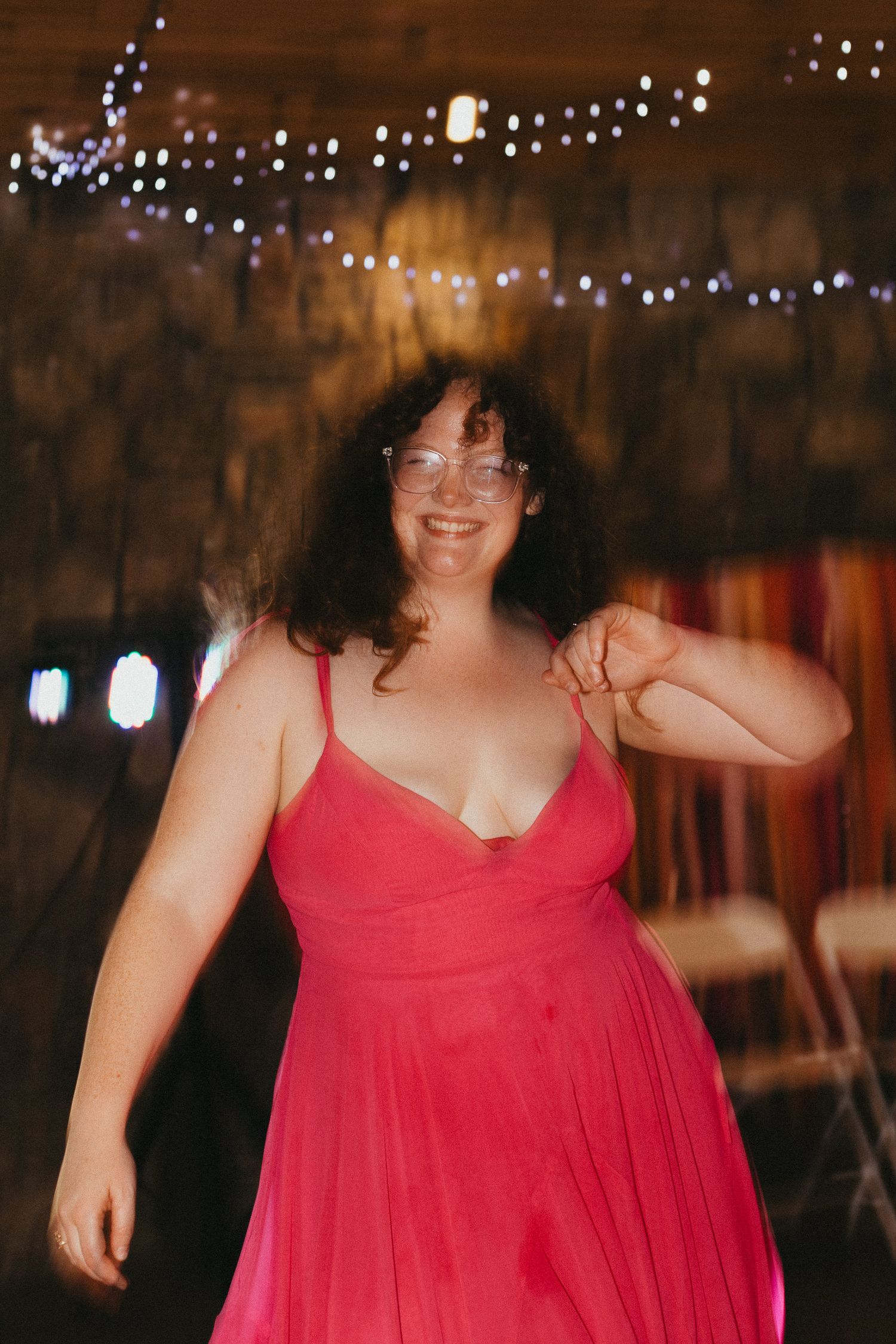 Woman in pink dress dancing joyfully at a wedding reception with fairy lights in the background.