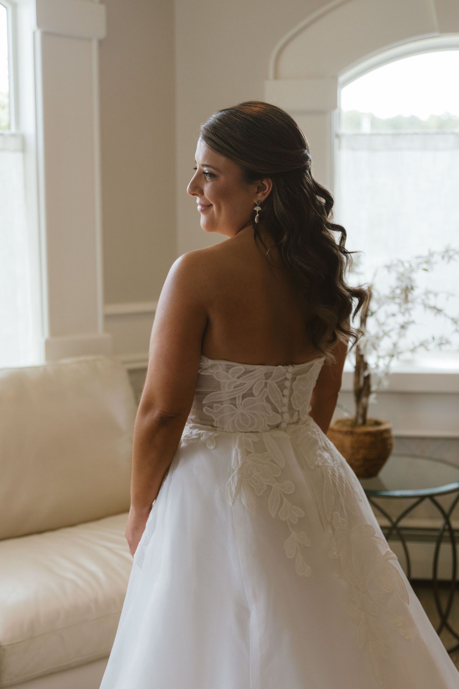 Bride in strapless white lace wedding gown looking over shoulder, smiling near bright windows in elegant room.