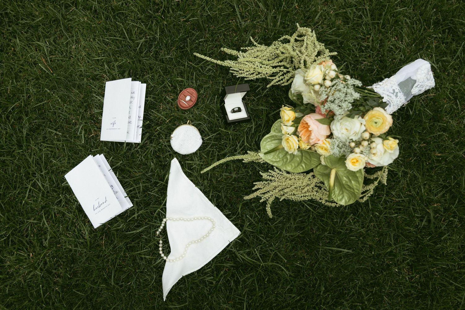 Flat lay of wedding accessories on green grass, including bouquet, rings, vows, candle, and white handkerchief.
