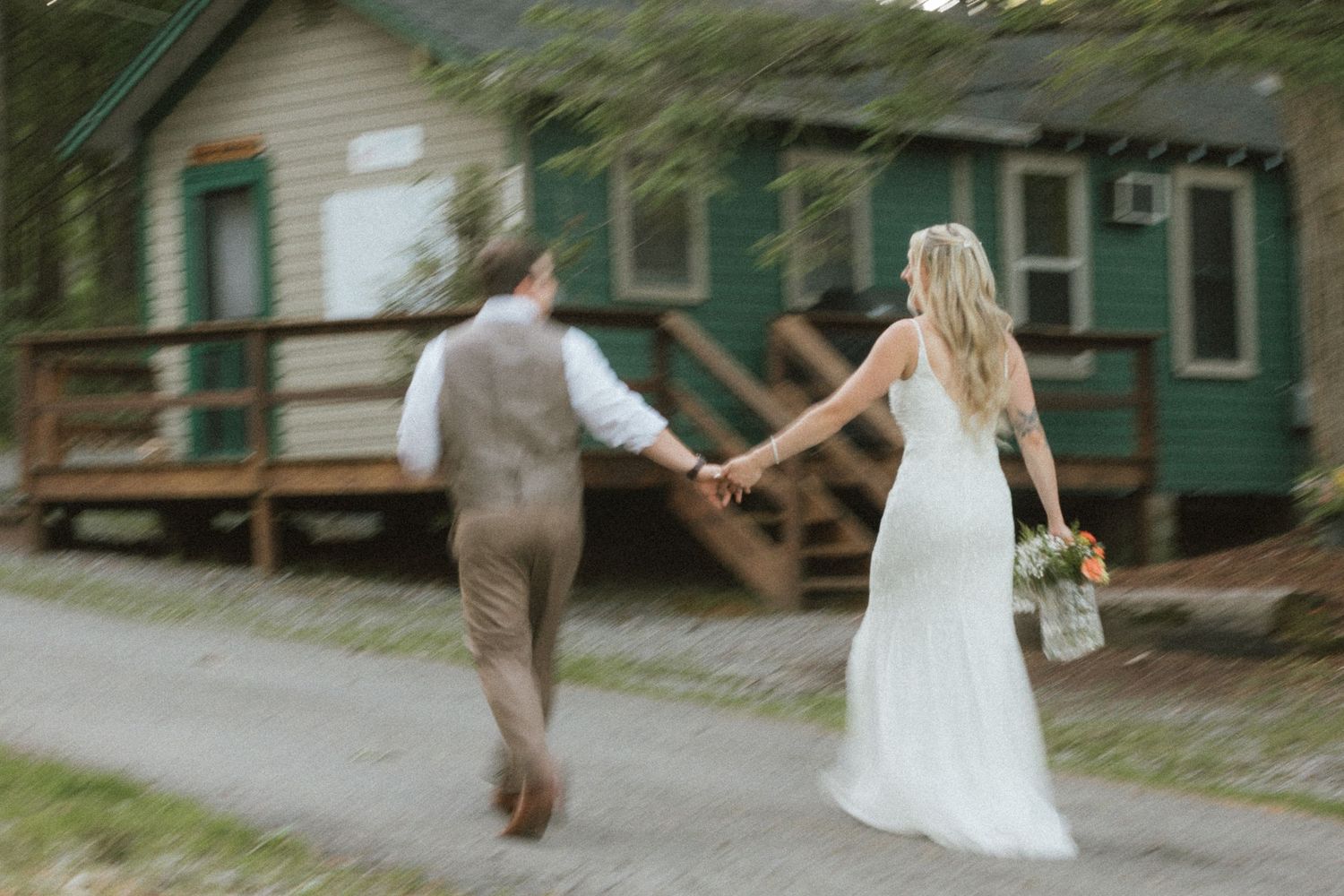 Bride and groom holding hands walking past a green cabin at their outdoor wedding.