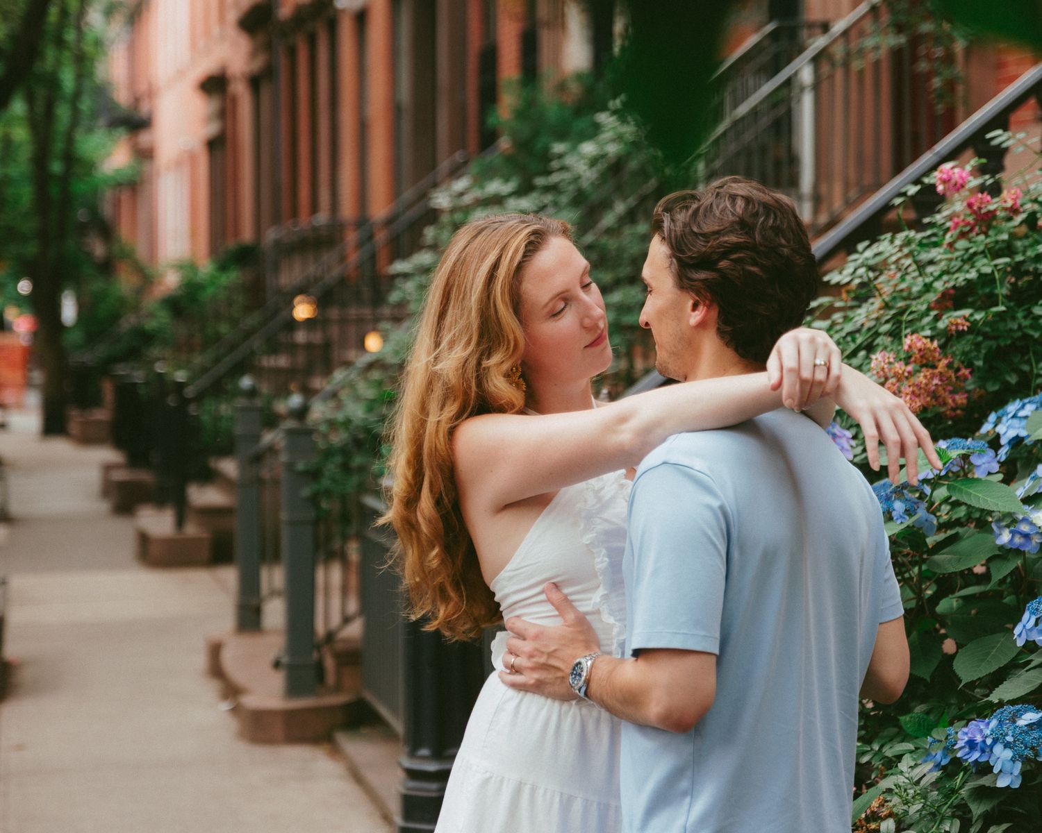 Couple embracing on a tree-lined urban sidewalk with brownstone buildings and blooming hydrangeas.