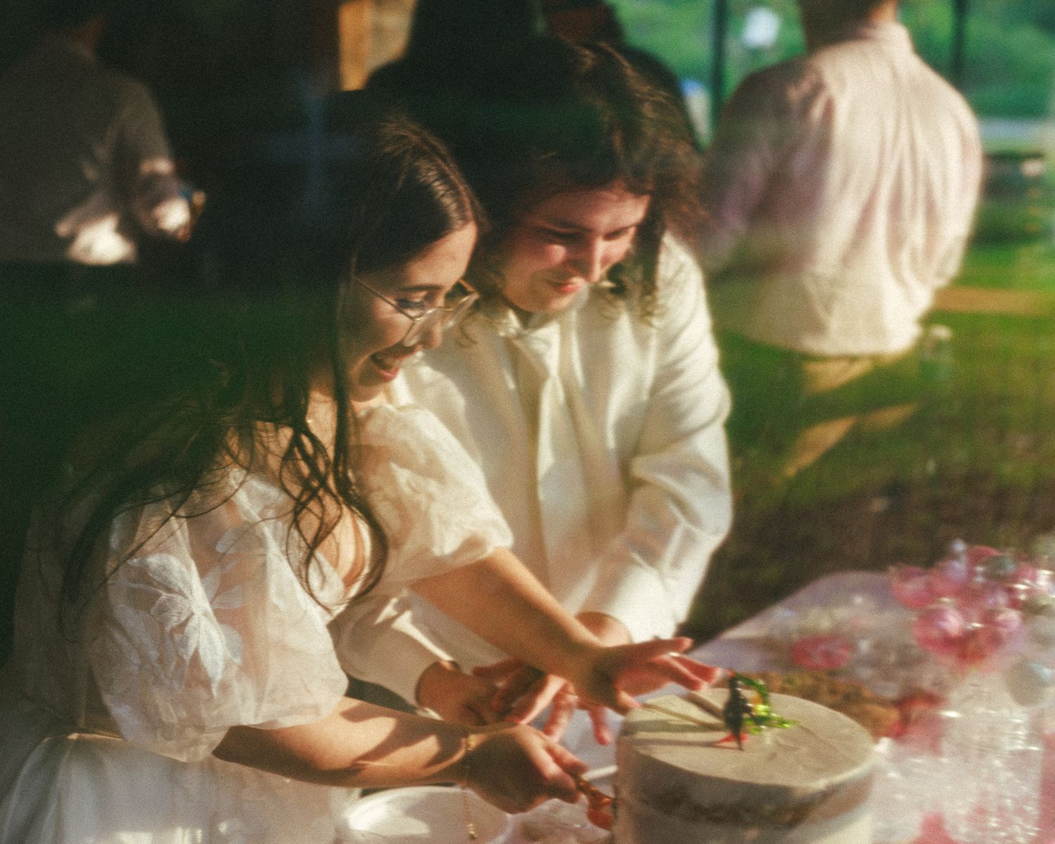Newlyweds cutting their wedding cake together outdoors in matching white outfits at evening reception.