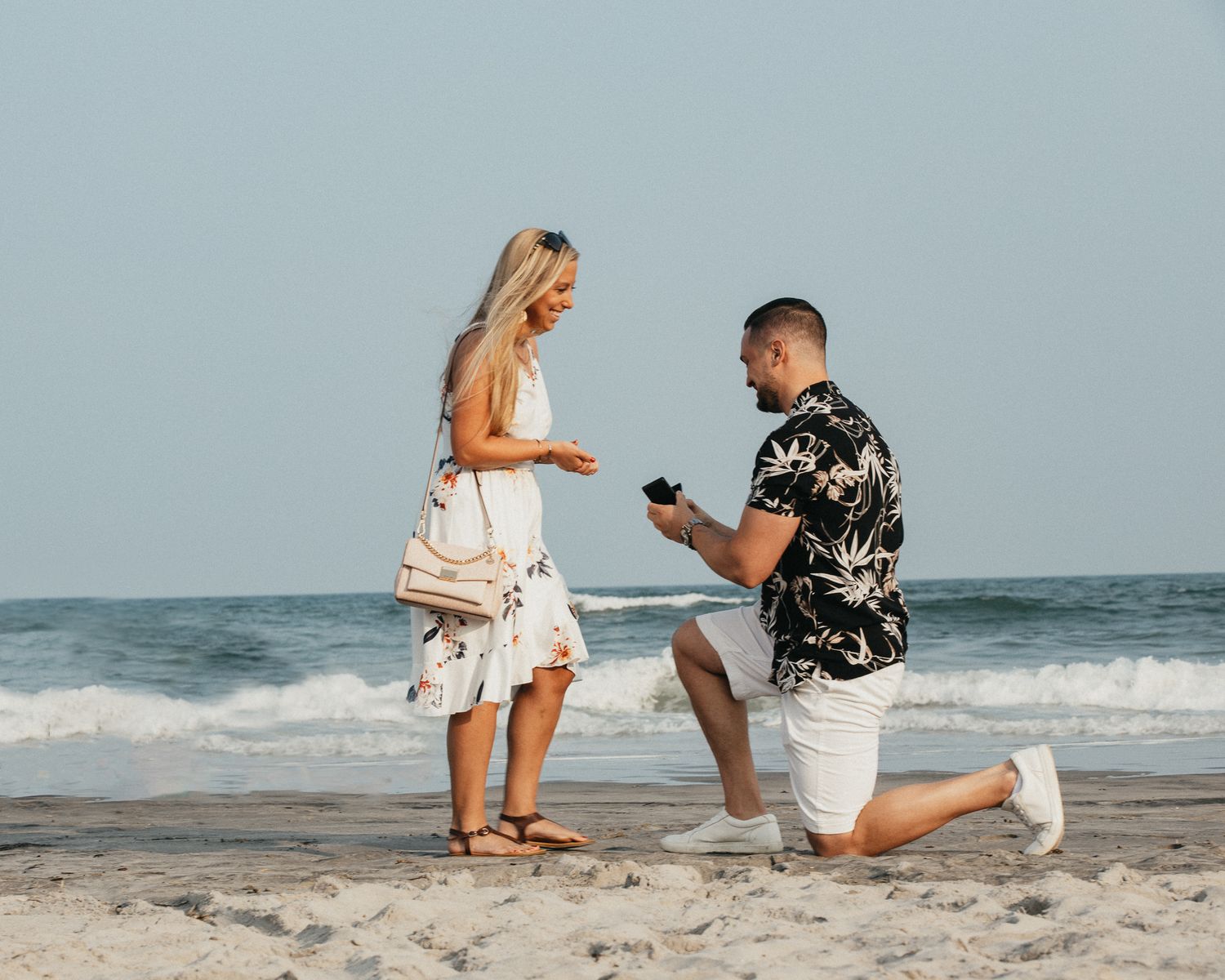 Romantic beach marriage proposal with man kneeling before blonde woman near ocean waves.