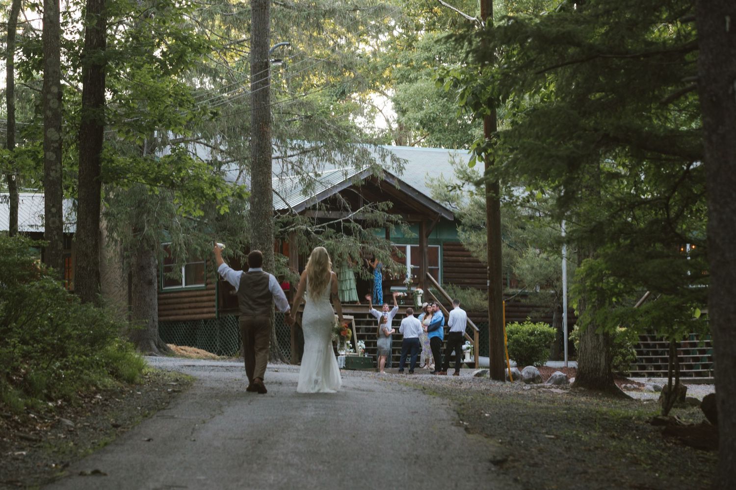 Bride and groom walk hand-in-hand down a forest path toward a rustic log cabin wedding venue.