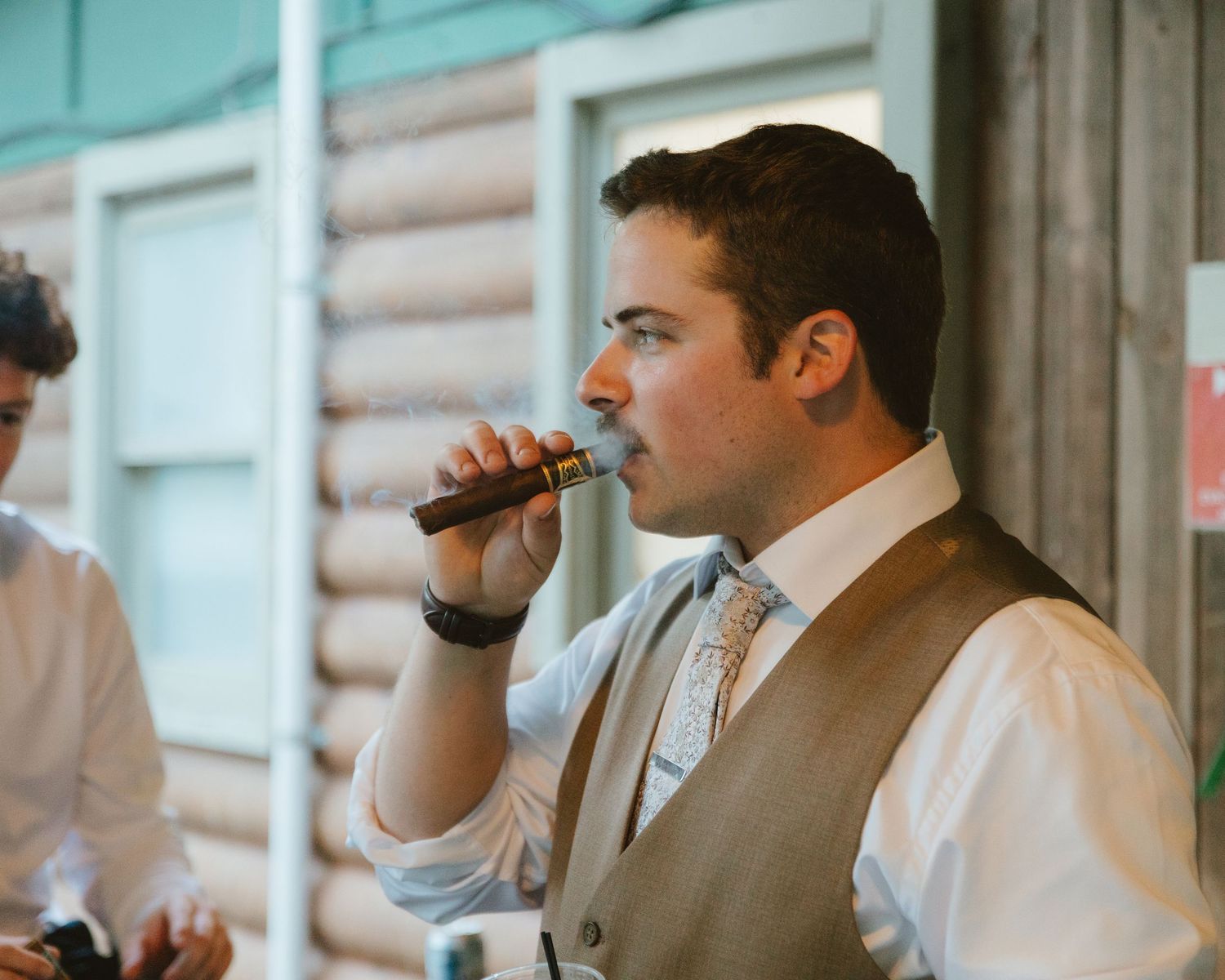 Man in vest and tie smoking a cigar at a rustic log cabin wedding reception.