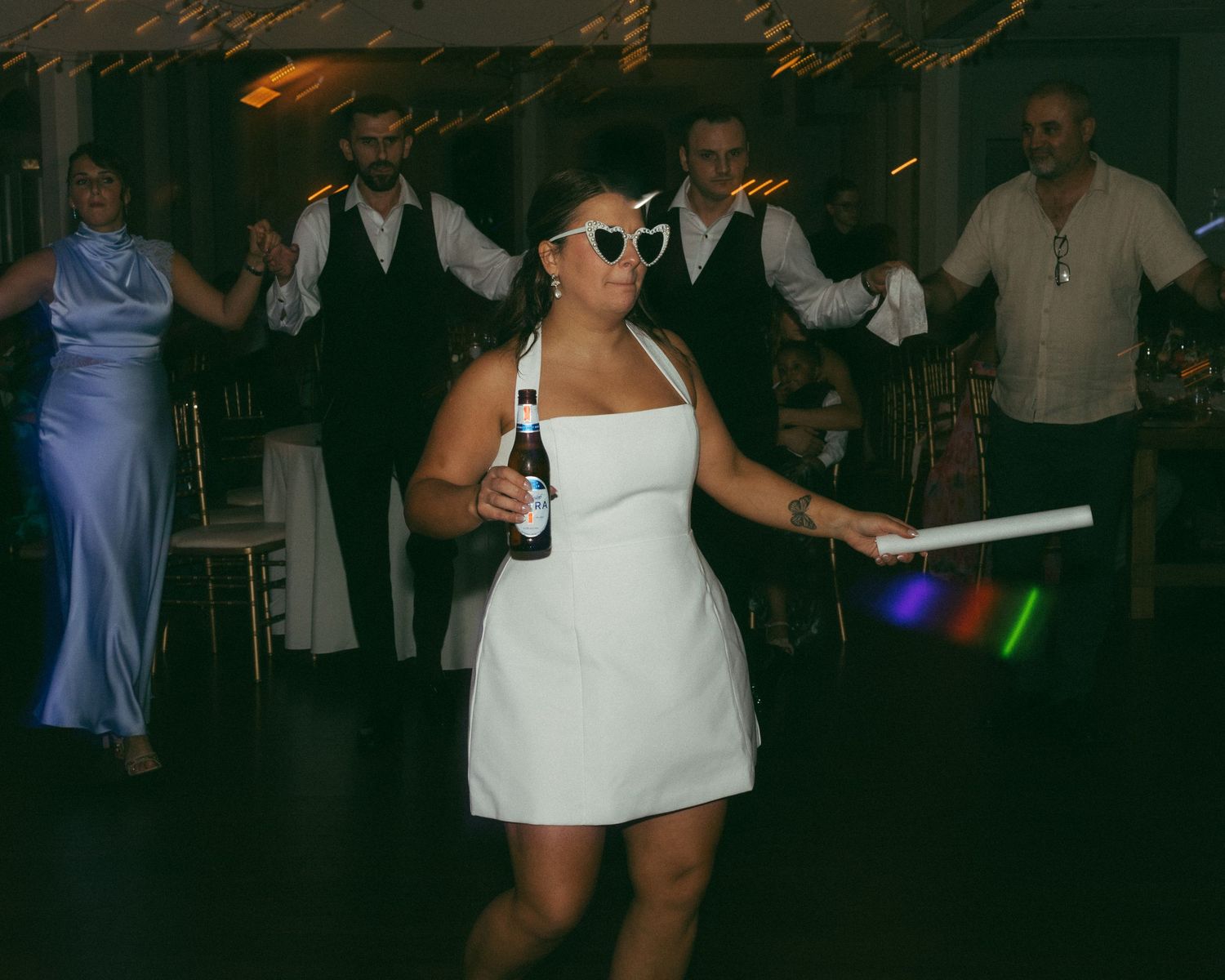 Bride in white dress and heart sunglasses dancing with glow sticks at a wedding reception party.