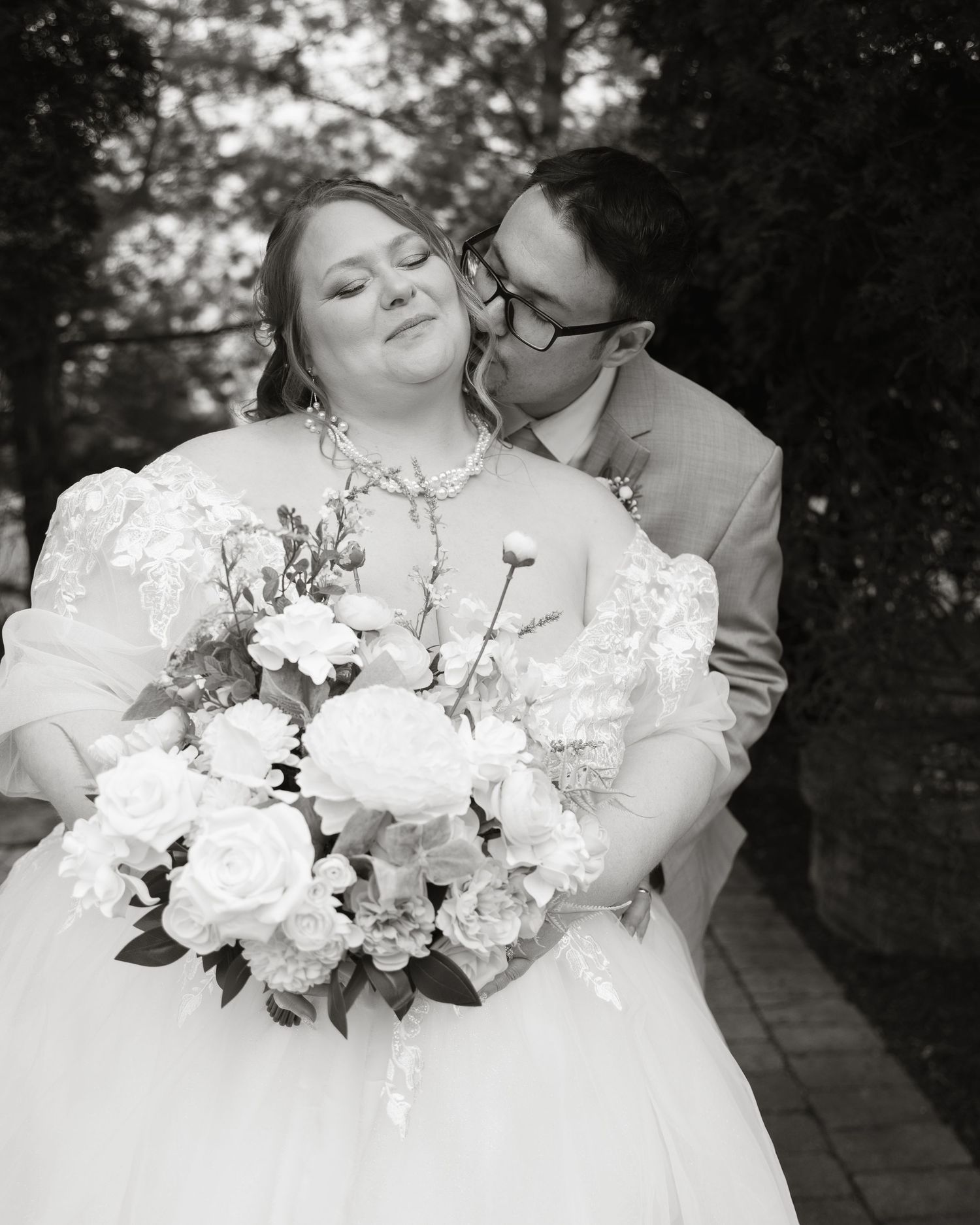 Black and white wedding photo of bride holding bouquet while groom kisses her cheek outdoors.