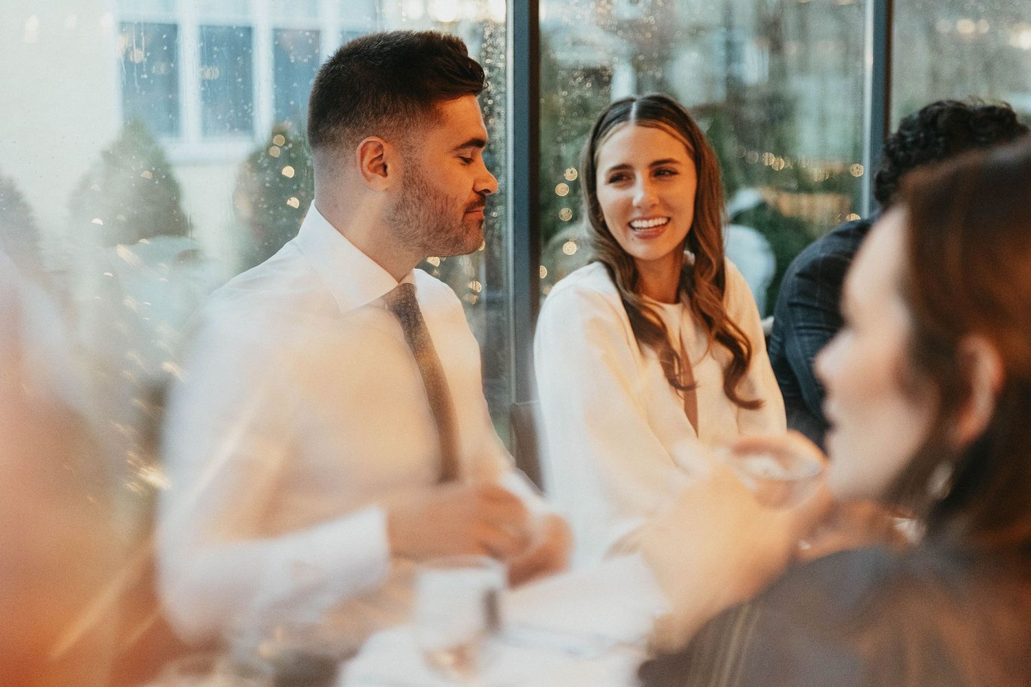 Smiling bride in white dress sits beside groom in white shirt at wedding reception table.