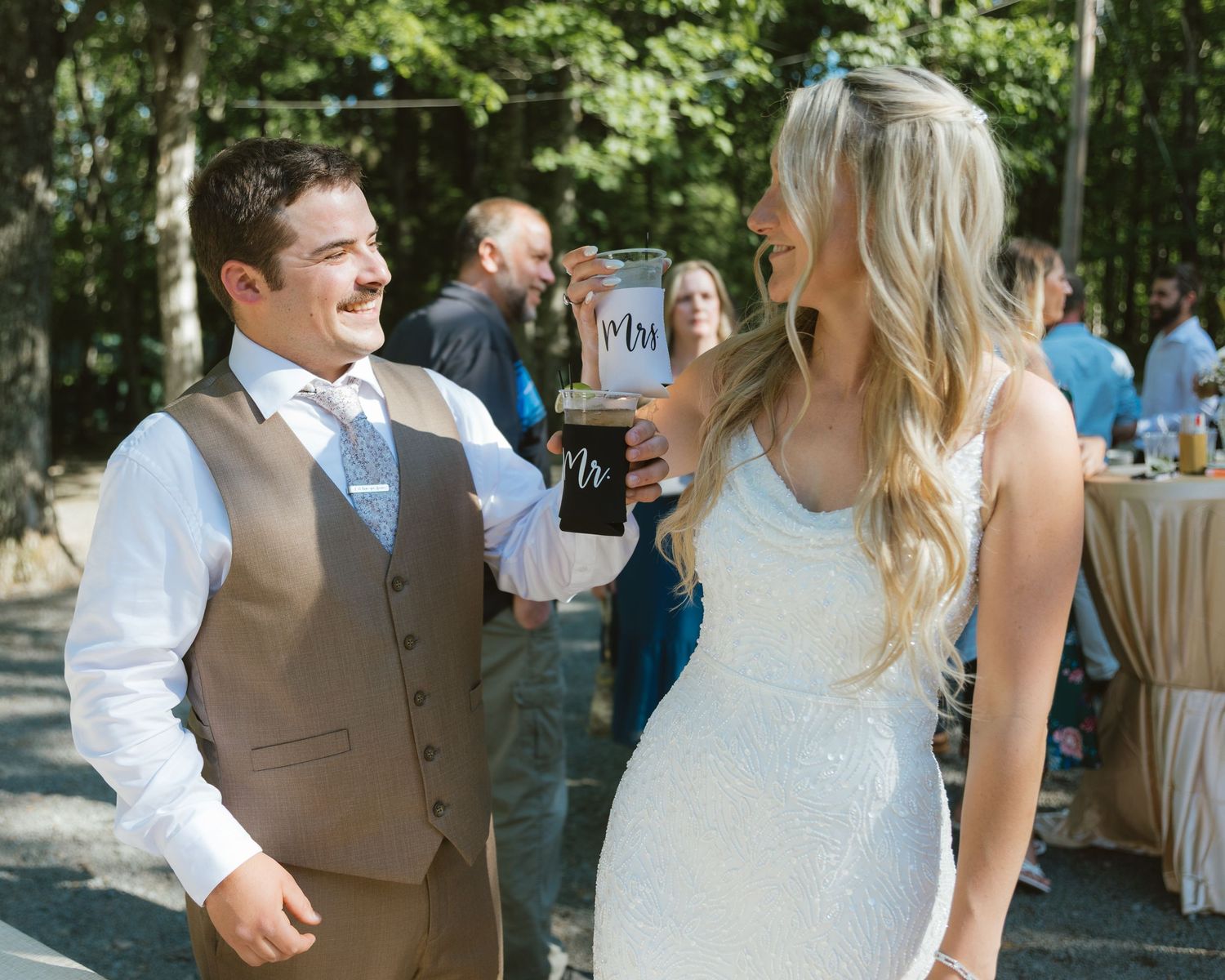 Bride and groom smiling at each other during outdoor wedding reception, holding personalized Mr and Mrs cups.