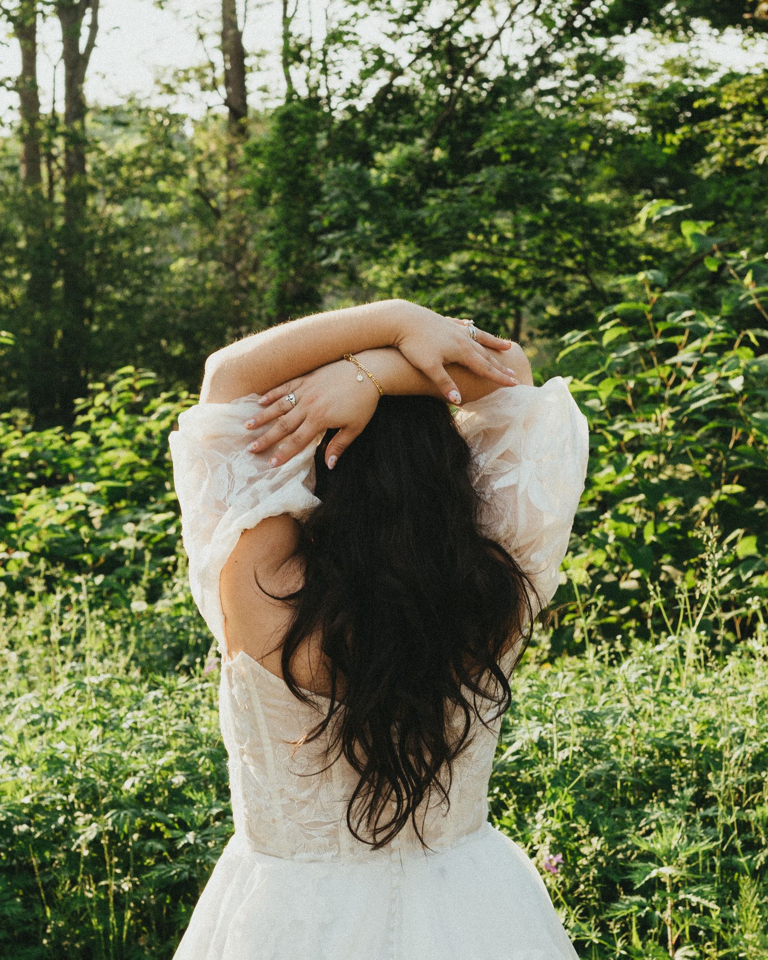 A woman with long dark hair in a white lace dress stands with arms raised in a lush green forest.