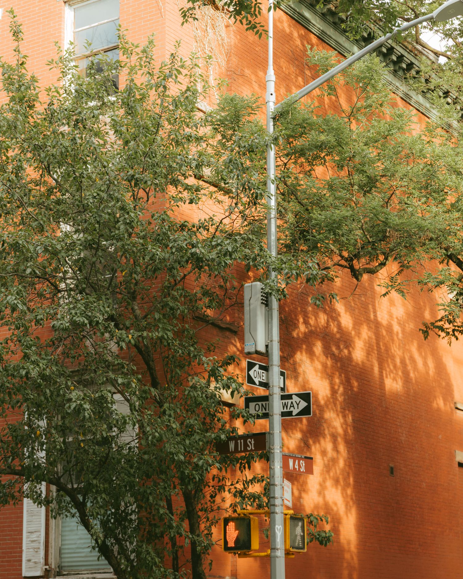 Street signs on a pole at a NYC intersection with lush green trees against a warm red-brick building facade.