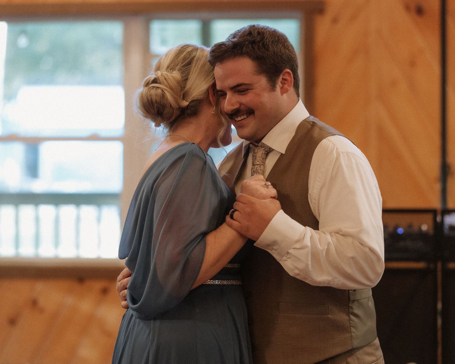 Couple sharing a romantic dance in a rustic wooden venue, man in vest and tie smiling at woman in blue dress.